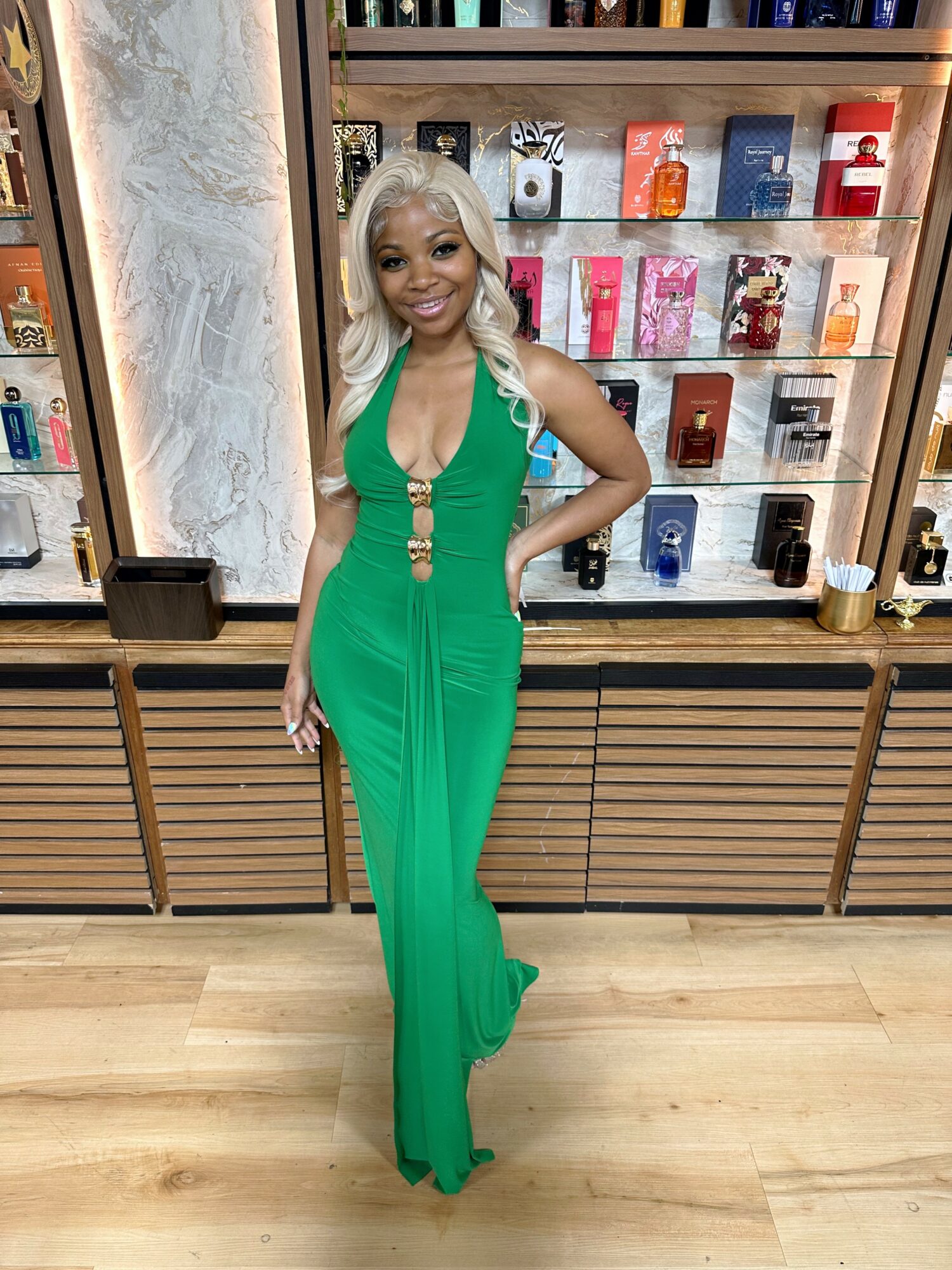 Woman in green jumpsuit standing in front of a display shelf with colorful items, smiling, with blonde hair and hand on hip.