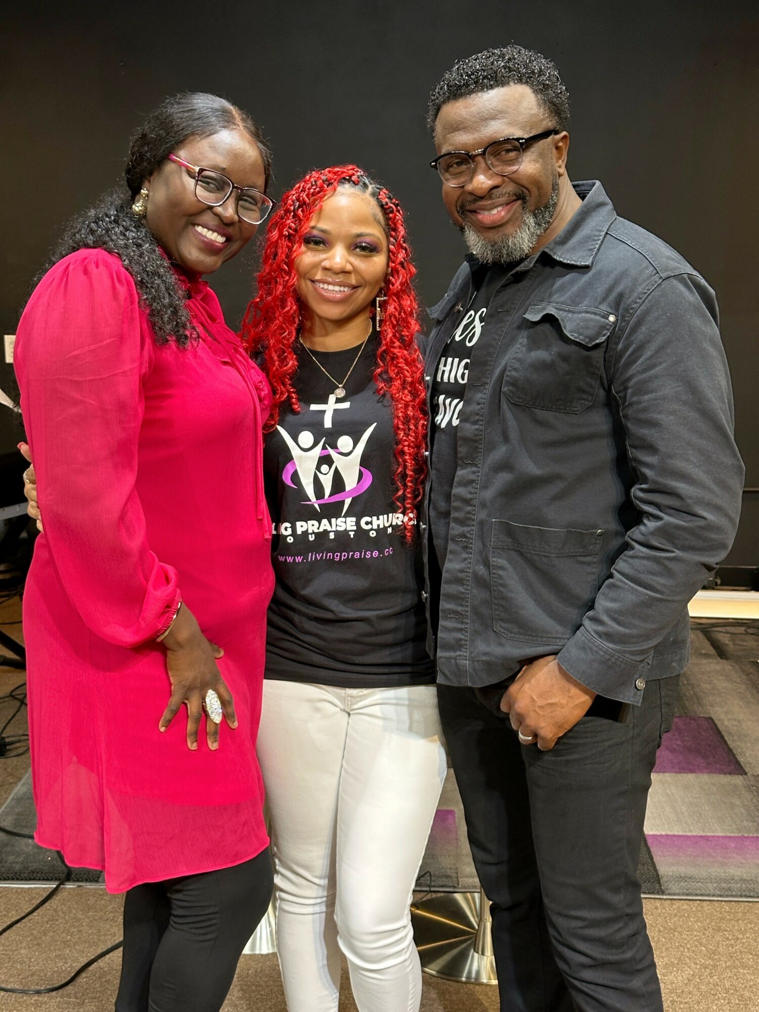 Three people smiling and standing together indoors, two women and one man, with a dark background.