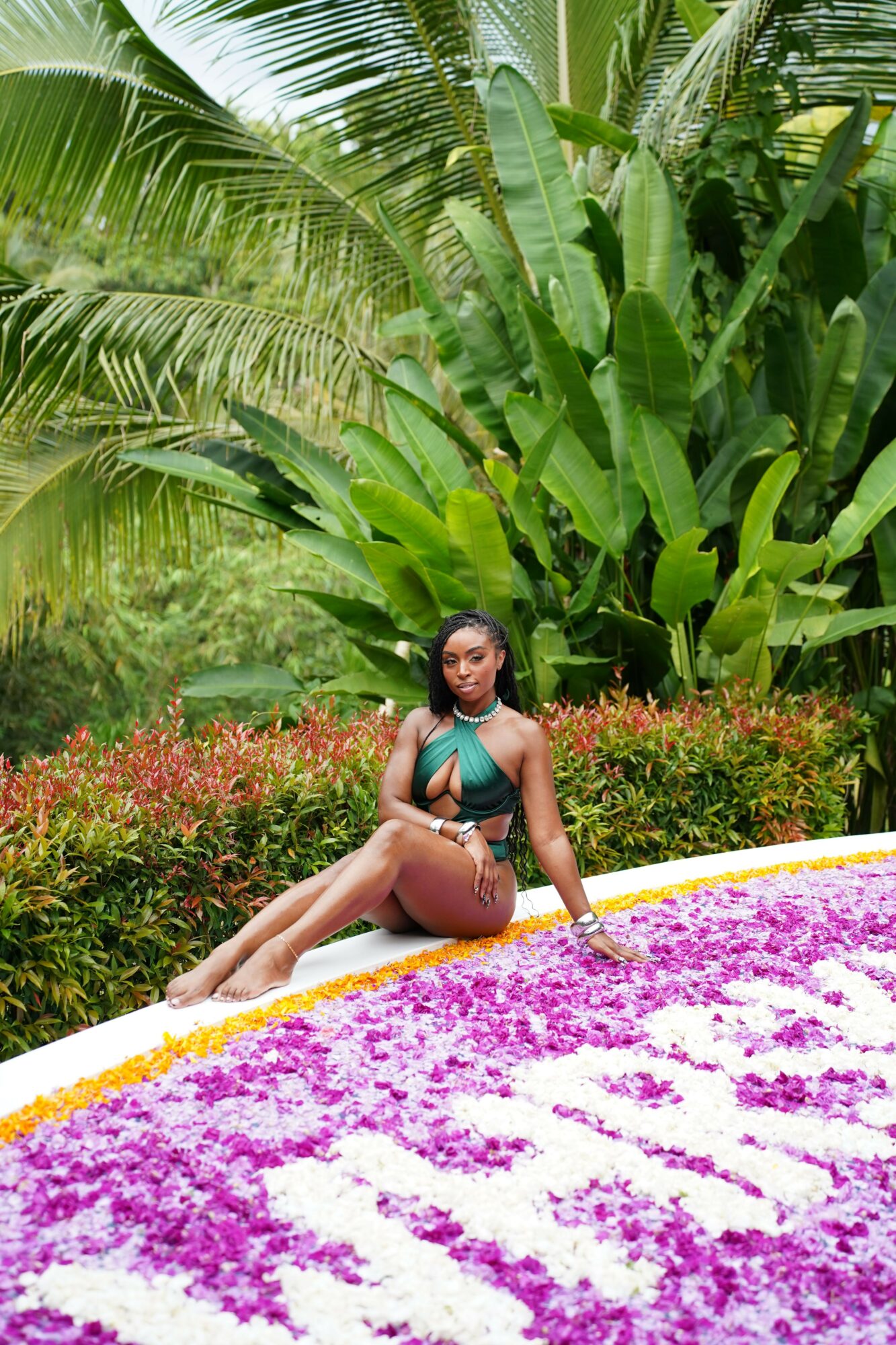 Woman in swimsuit sitting on a flower bed with green tropical plants in the background.
