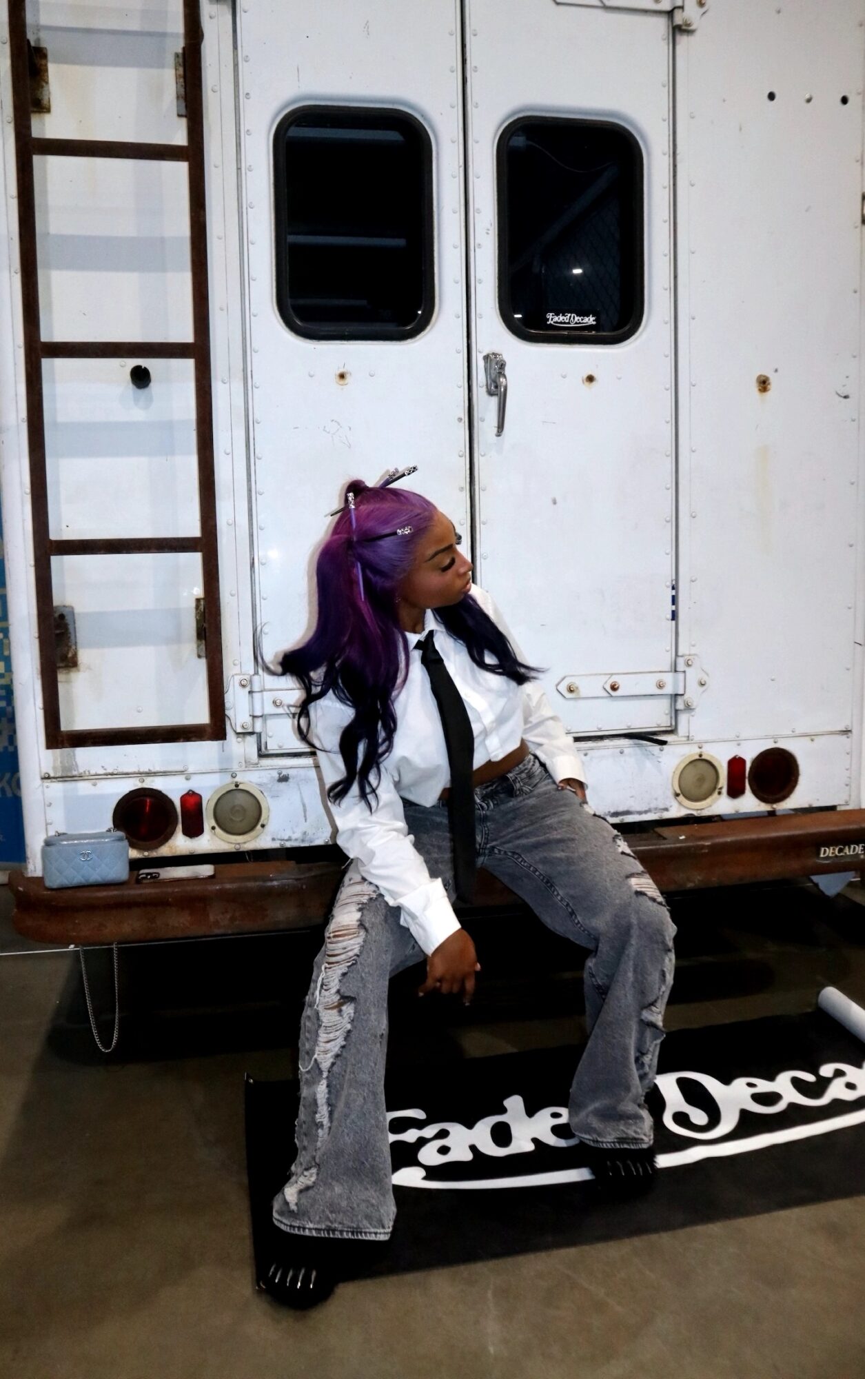 Person with purple hair and white jacket sitting on a black and white sign in front of a white trailer door.