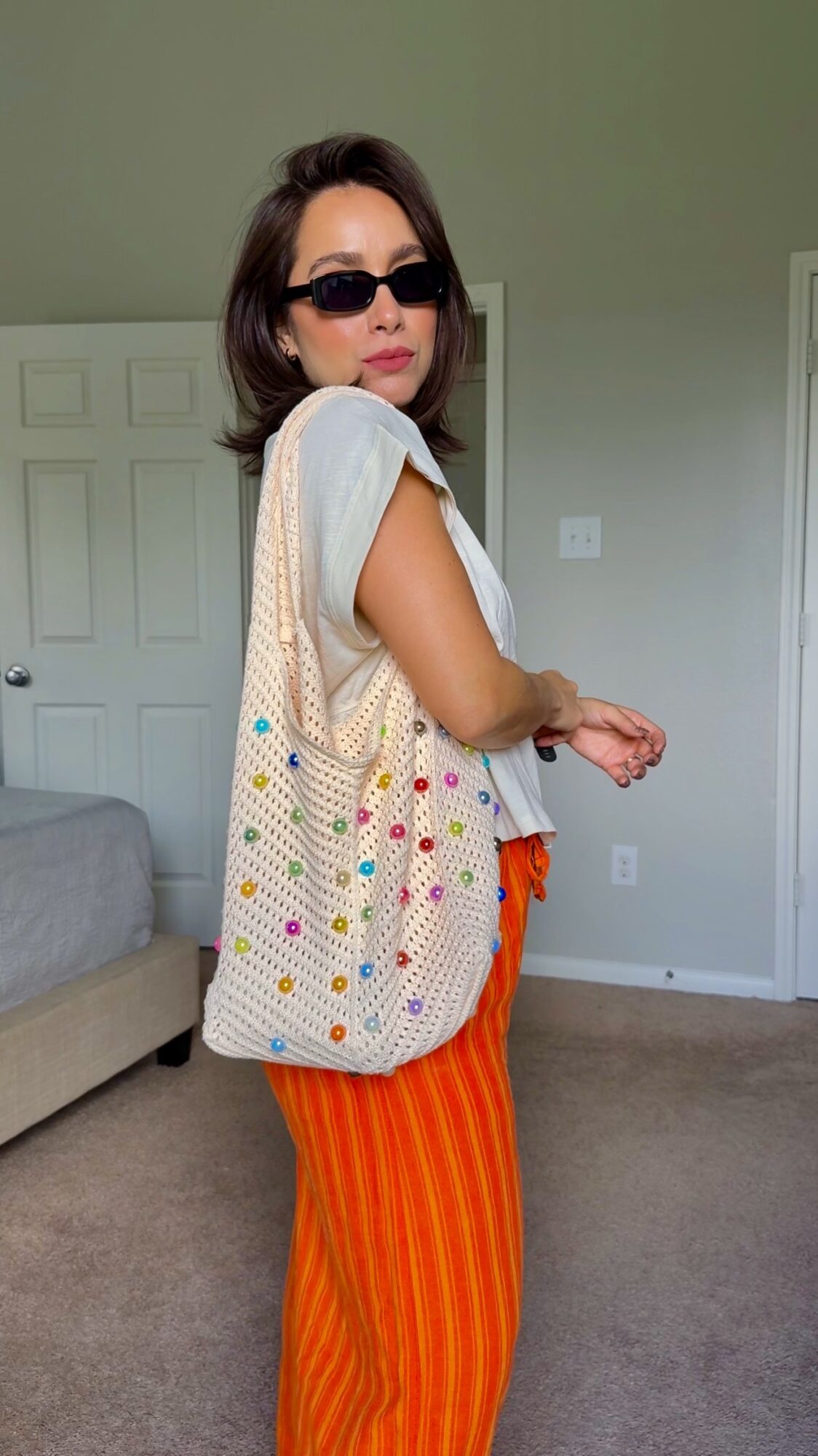 Woman with sunglasses carrying a large polka dot bag, wearing orange striped pants and a light top, standing indoors.