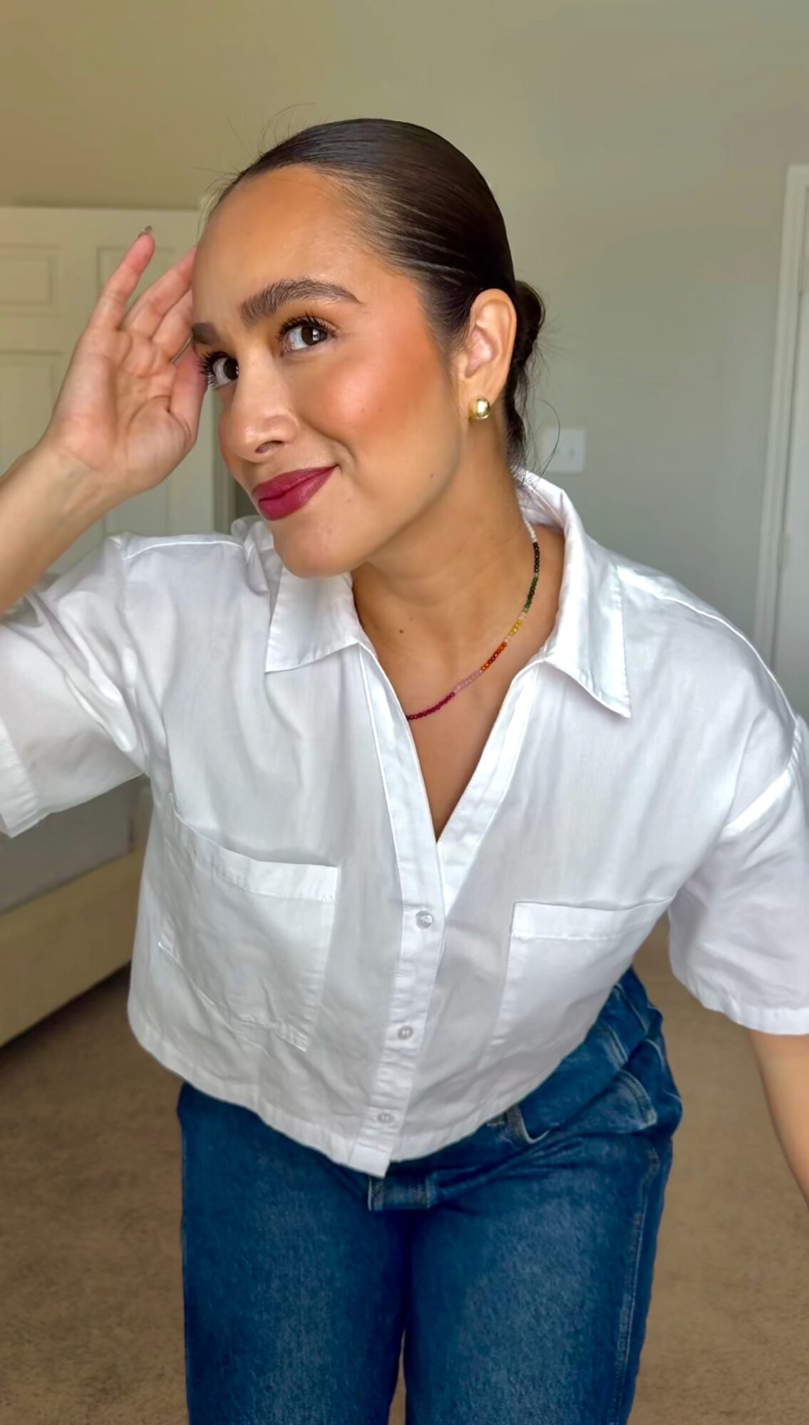 Woman with dark hair in a bun, wearing a white shirt and blue jeans, smiling and touching her forehead.