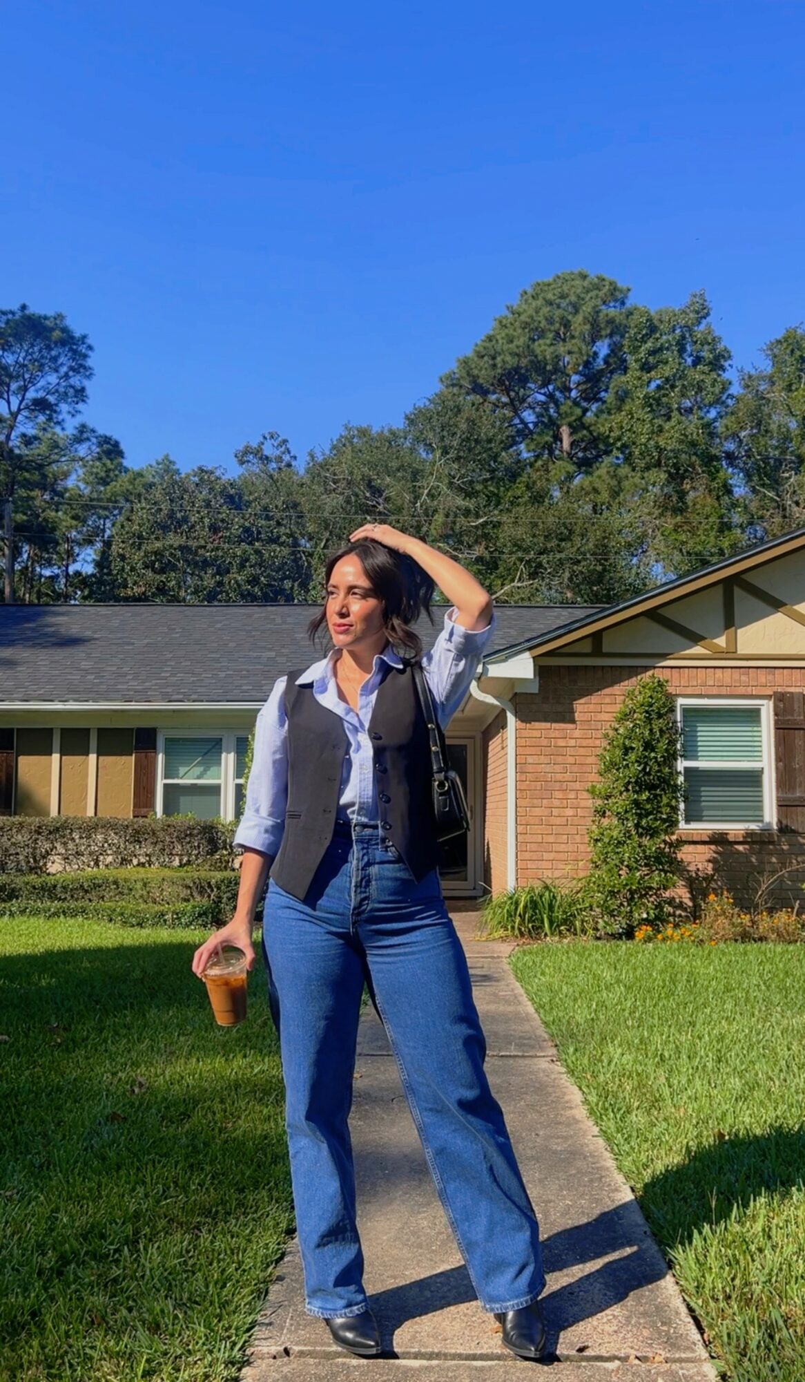 Woman standing on sidewalk in front of house, holding a drink, with trees and blue sky in background.