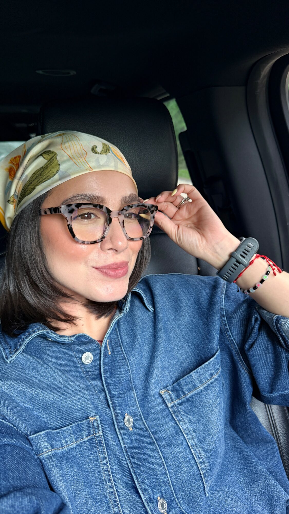 Woman wearing glasses and a headscarf, sitting in a vehicle, adjusting her glasses, with a denim shirt and bracelets.