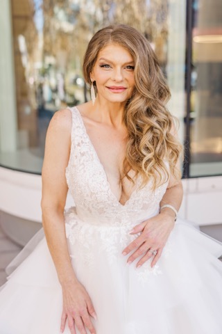 Woman with long wavy hair in a white dress outdoors, smiling, with trees and building reflections behind her.
