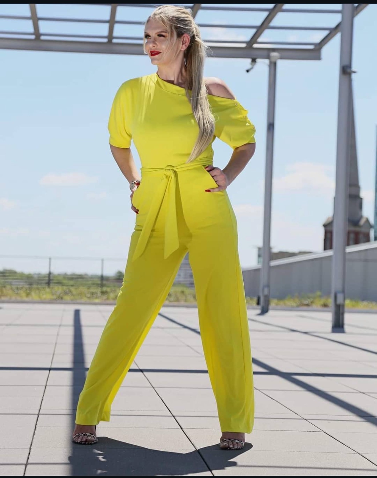Woman in yellow jumpsuit with off-shoulder sleeves standing outdoors on a tiled surface, with a modern structure and sky in background.