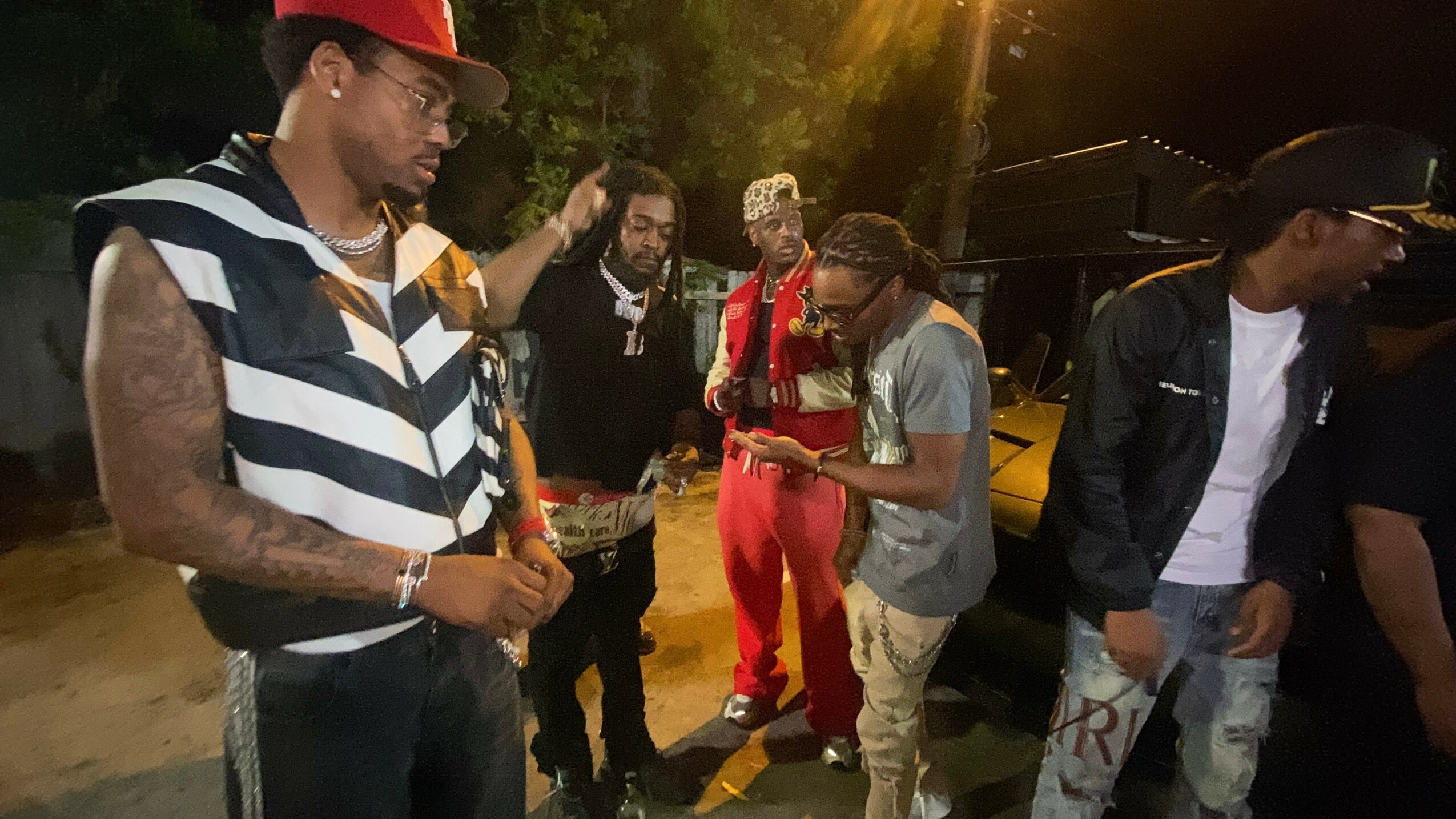 Group of six people standing outdoors at night, some looking at their phones, with trees and lights in the background.