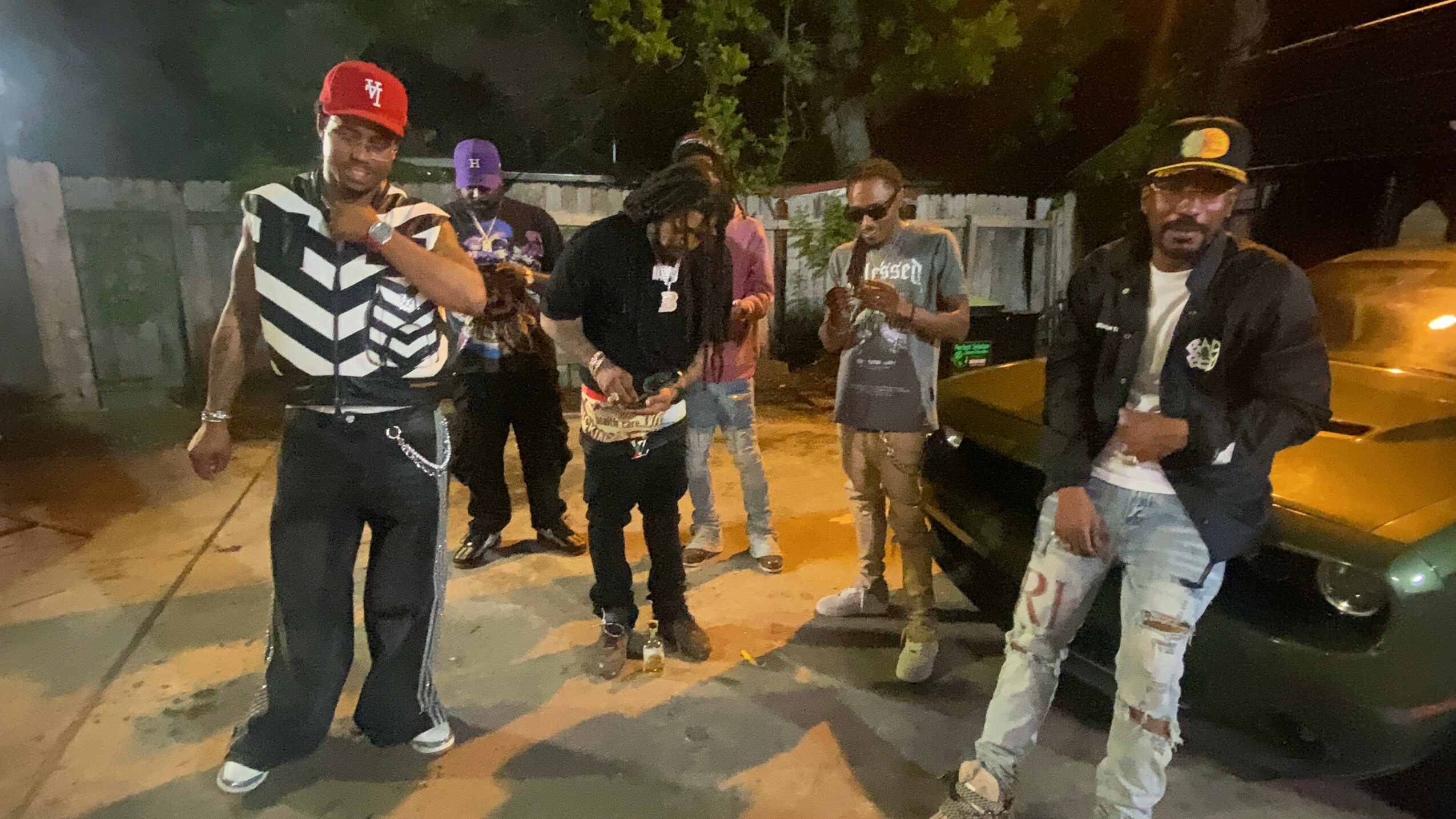 Group of people standing outdoors at night, some wearing casual clothing and sunglasses, with trees and a yellow vehicle in the background.