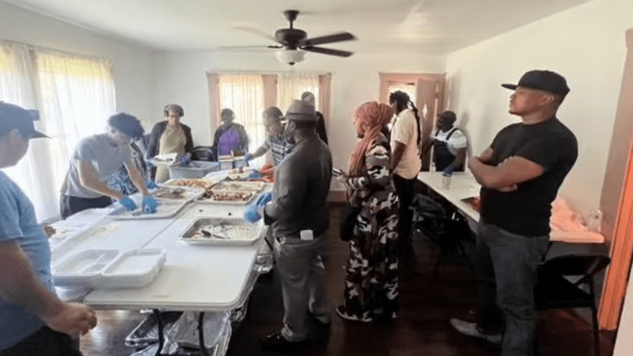 People gather around tables with food in a room with windows and a ceiling fan.