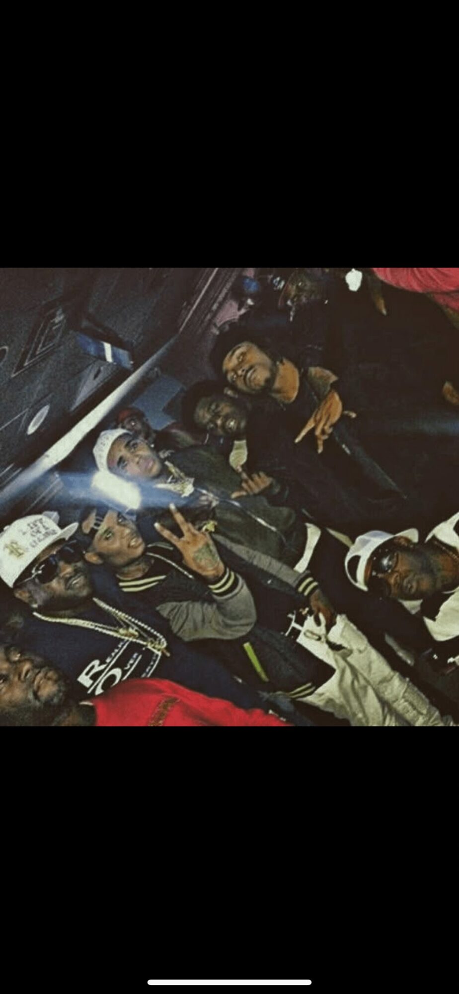 Group of people sitting together in a vehicle, some making peace signs, with dark clothing and accessories.