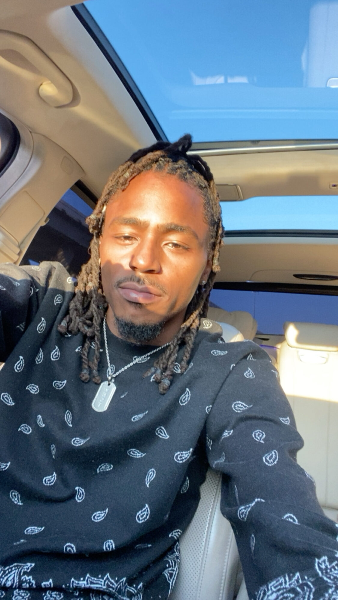 Young man with dreadlocks taking a selfie inside a car with a sunroof, blue sky visible outside.