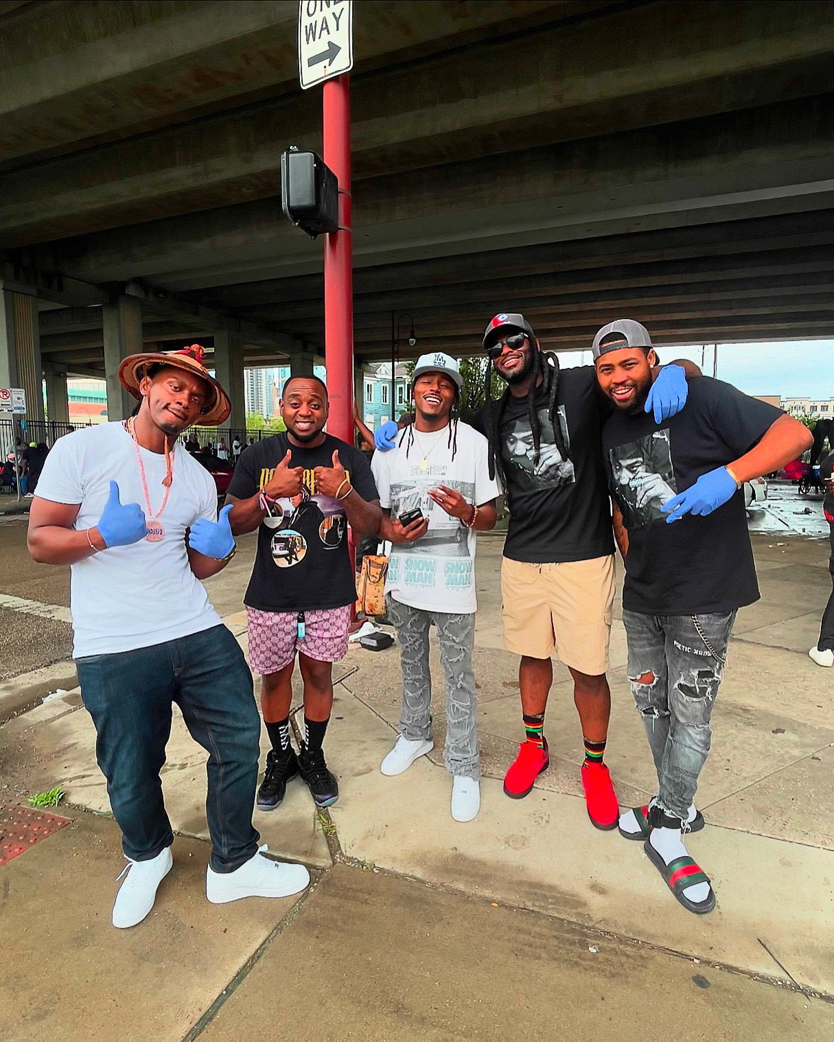 Group of five people standing outdoors under a bridge, smiling and giving thumbs up, with a red pole and street sign nearby.