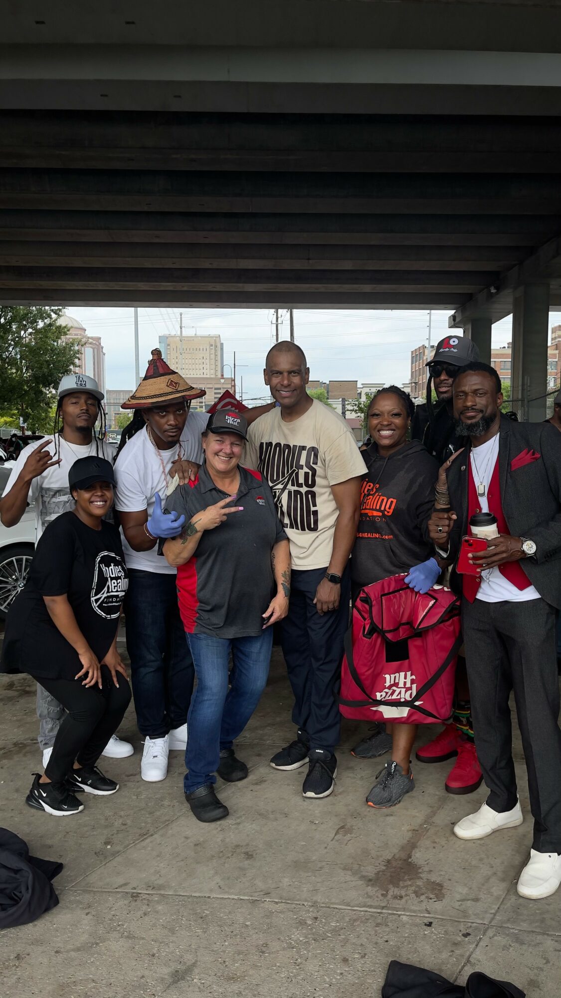 Group of people standing outdoors under a bridge, smiling, with trees and buildings in the background.