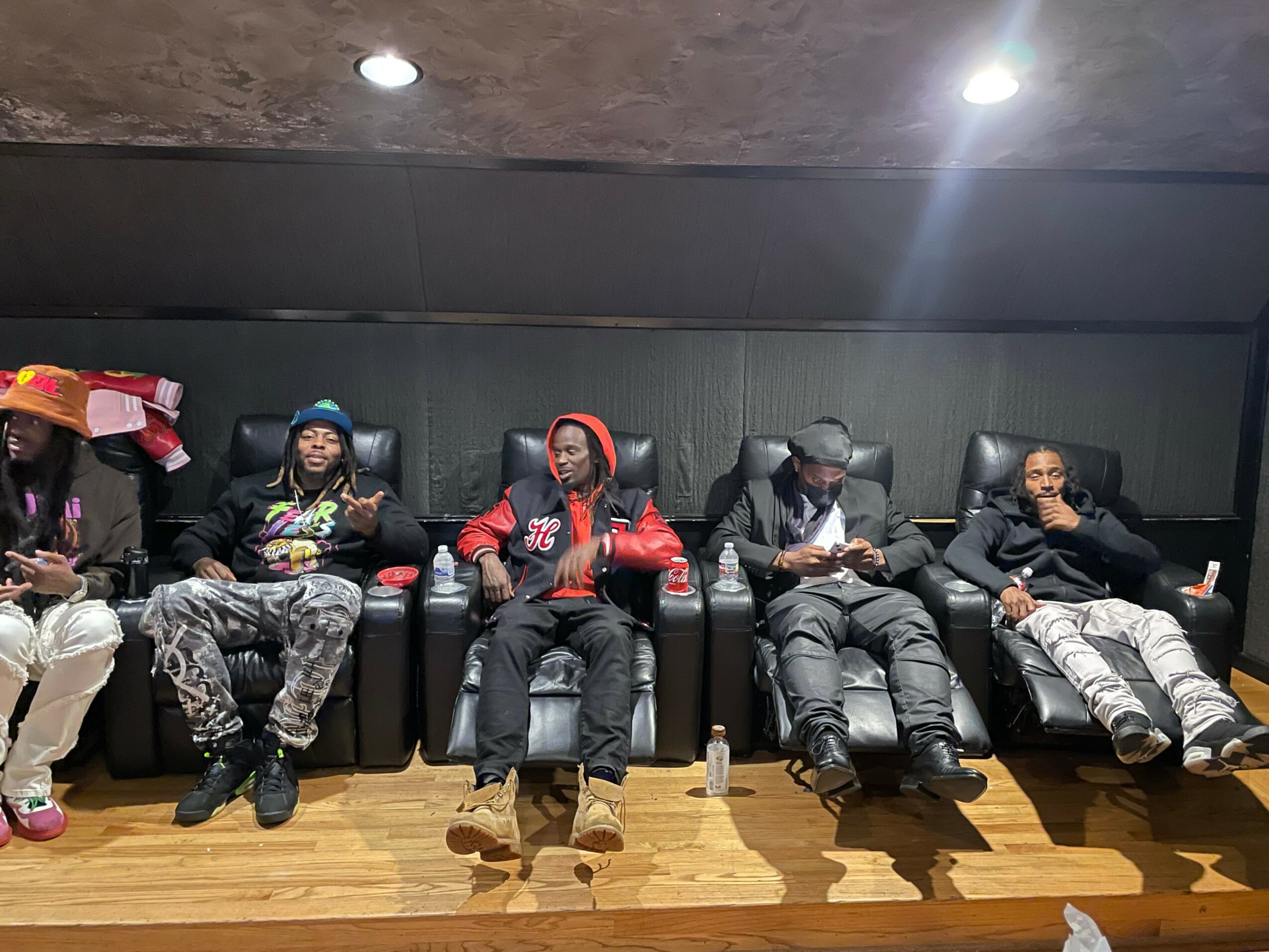 Five people sitting in black recliner chairs against a dark wall, some using phones, one with a hat, in a room with wooden floor.
