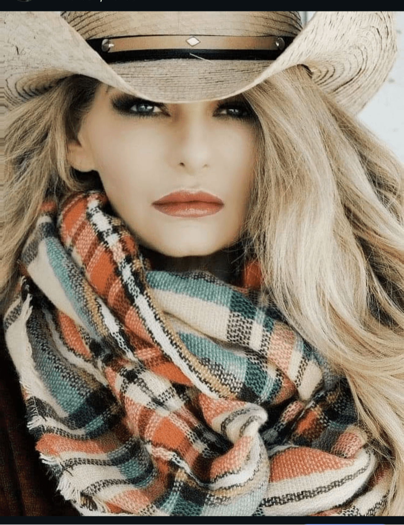 Woman wearing a wide-brimmed hat and a colorful plaid scarf, with long blonde hair and makeup.