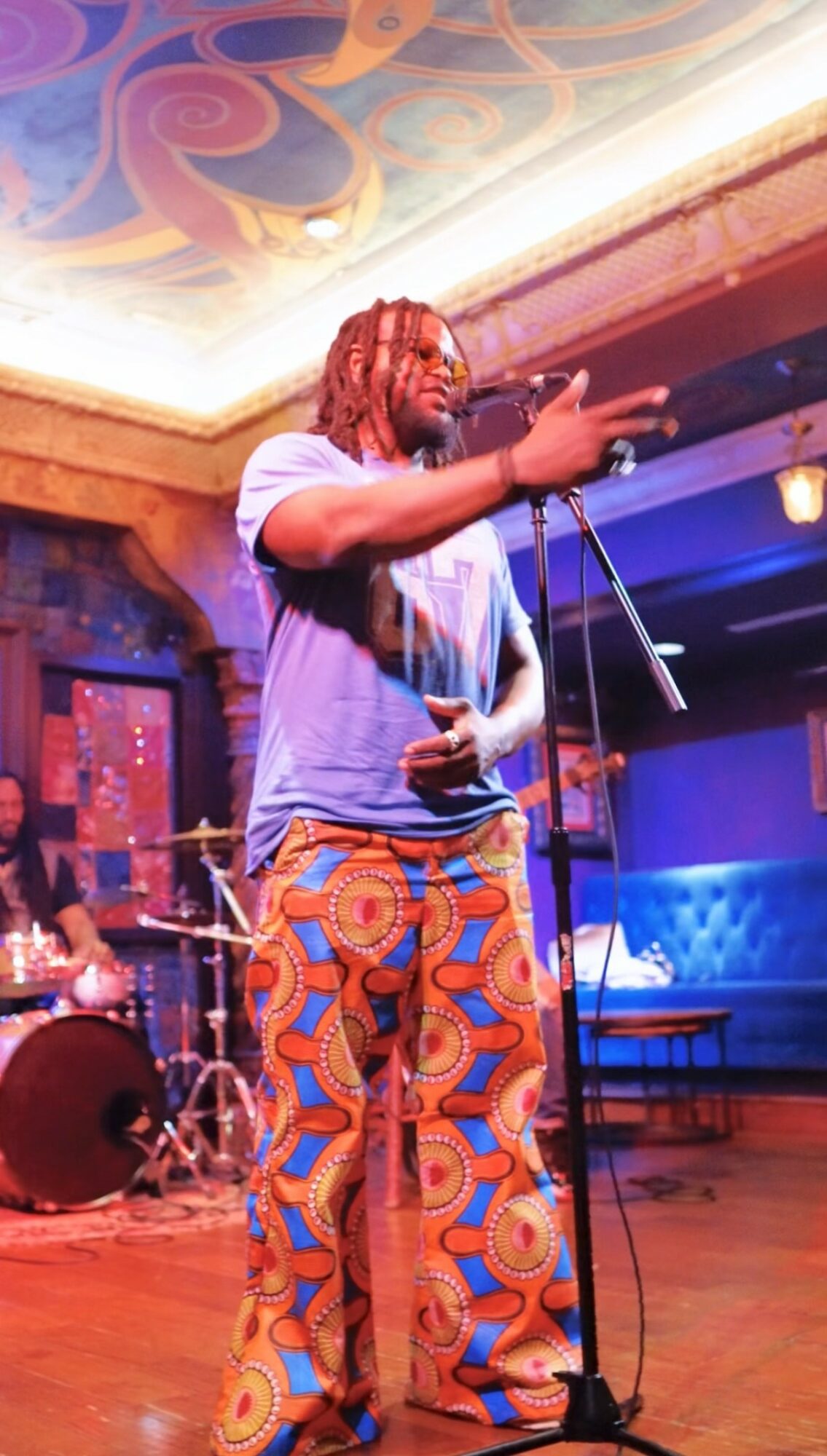 Performer with red dreadlocks singing into a microphone on stage, wearing colorful patterned pants and a gray T-shirt.