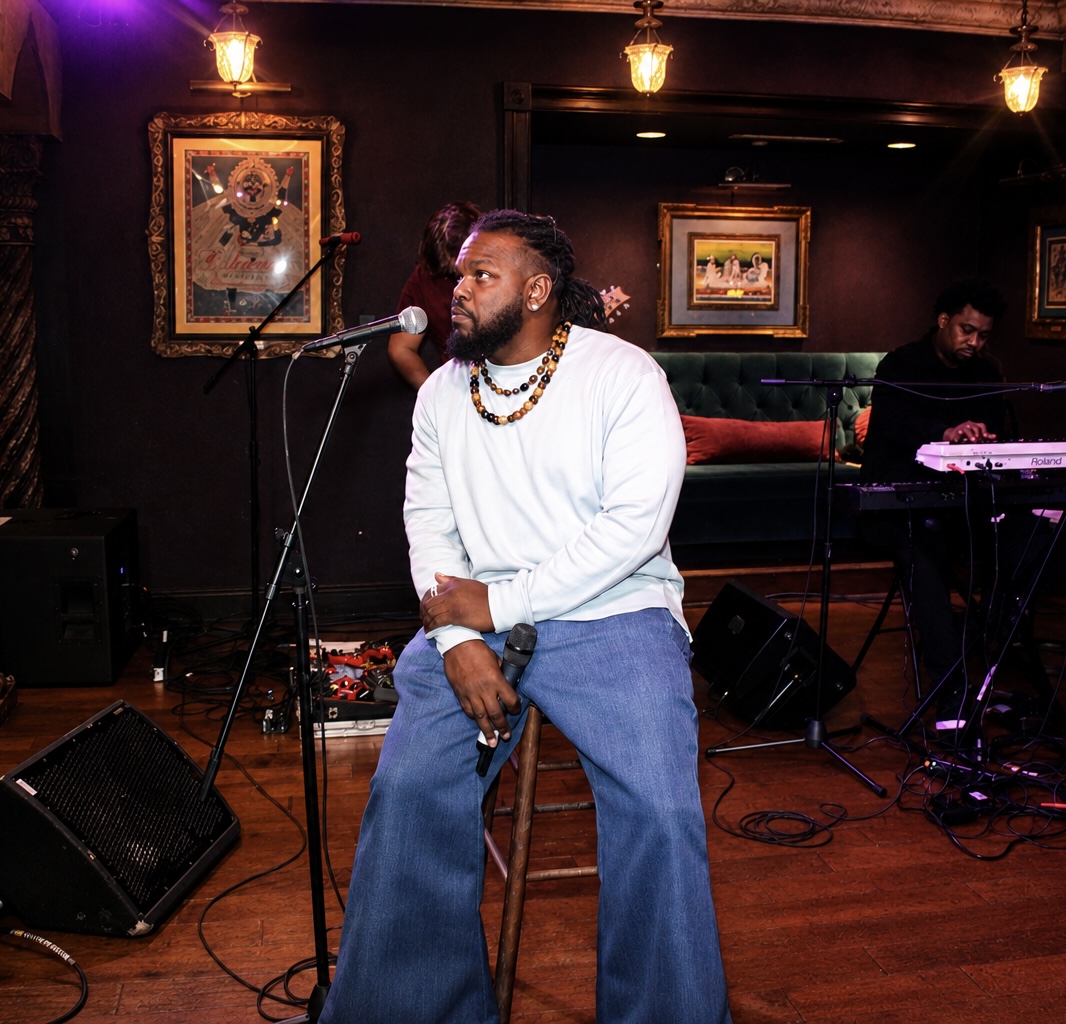 Man sitting on a stool with a microphone, performing on stage with a keyboard player in the background, framed artwork on dark walls.