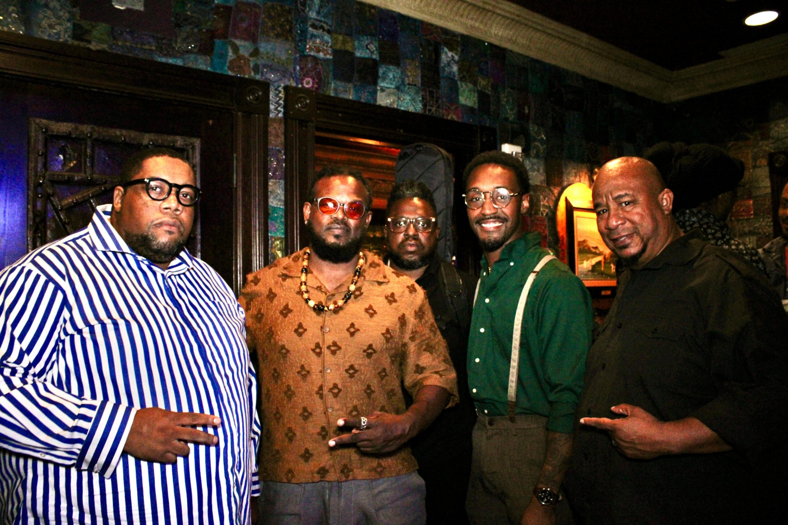 Five men standing together indoors, some wearing glasses, smiling, with dark walls and warm lighting in the background.