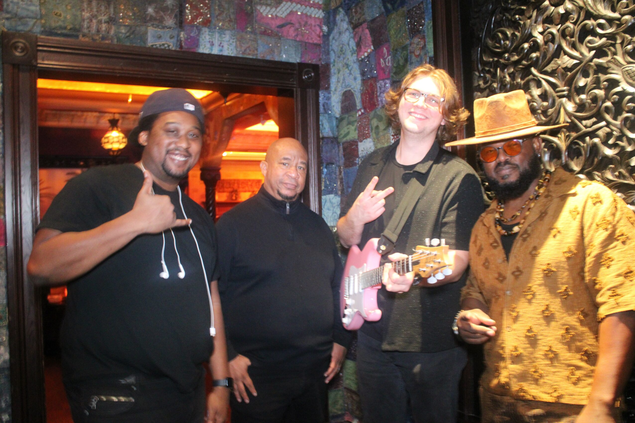 Four men standing together indoors, smiling, with decorative background and warm lighting.