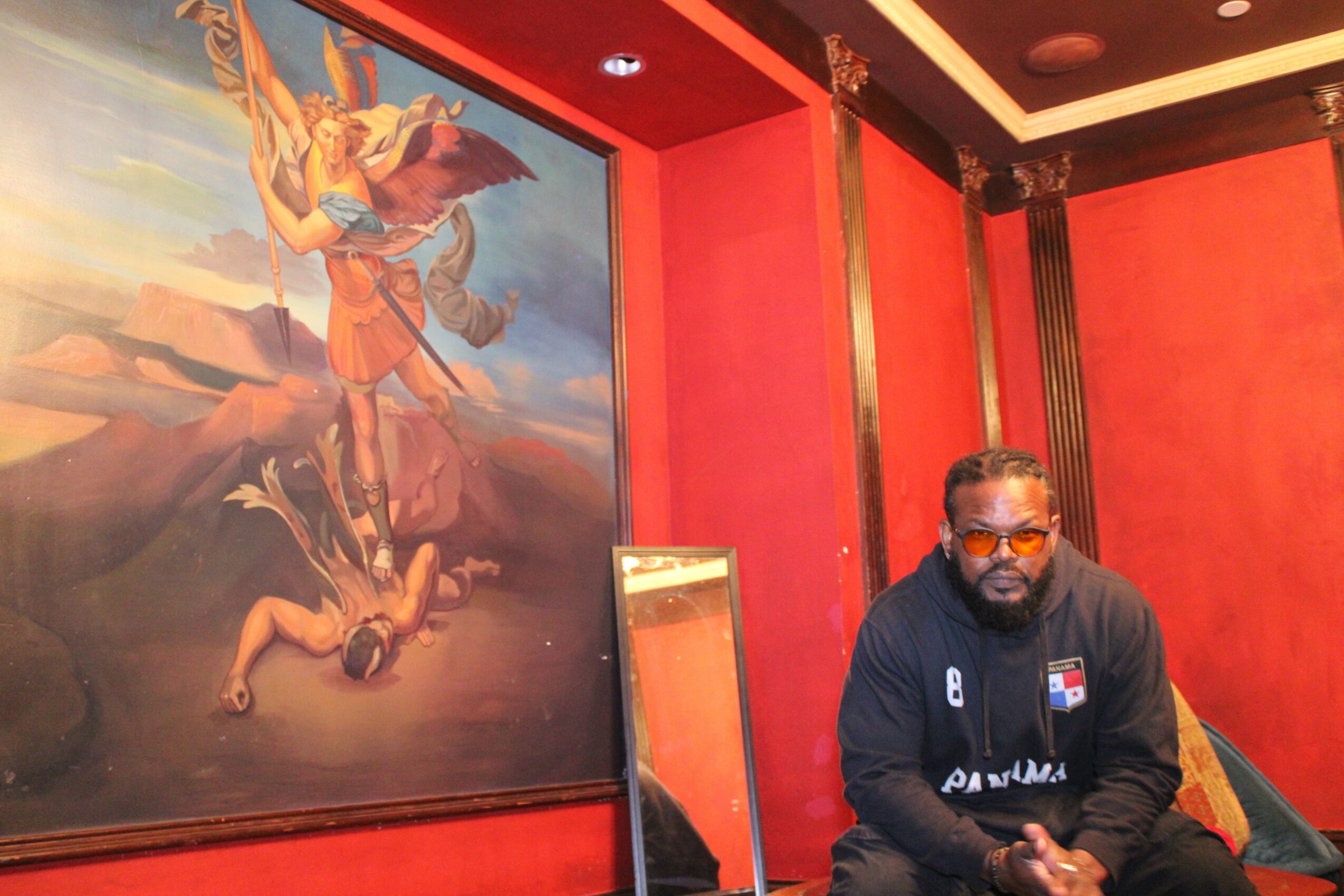 Man with glasses and beard sitting indoors with red walls and a large painting behind him.