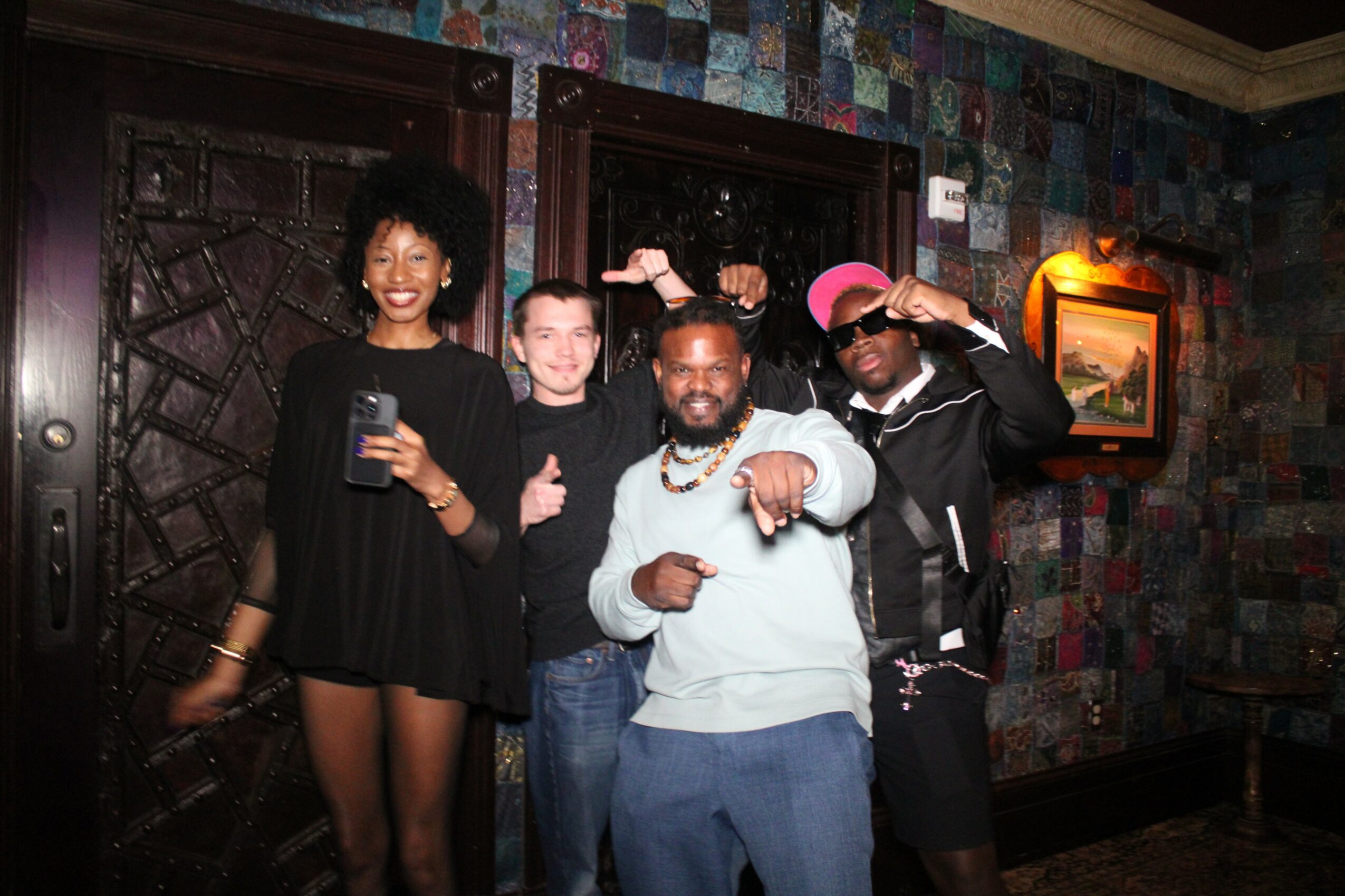 Group of five people smiling and posing indoors, with colorful wall background and framed picture, some making gestures.