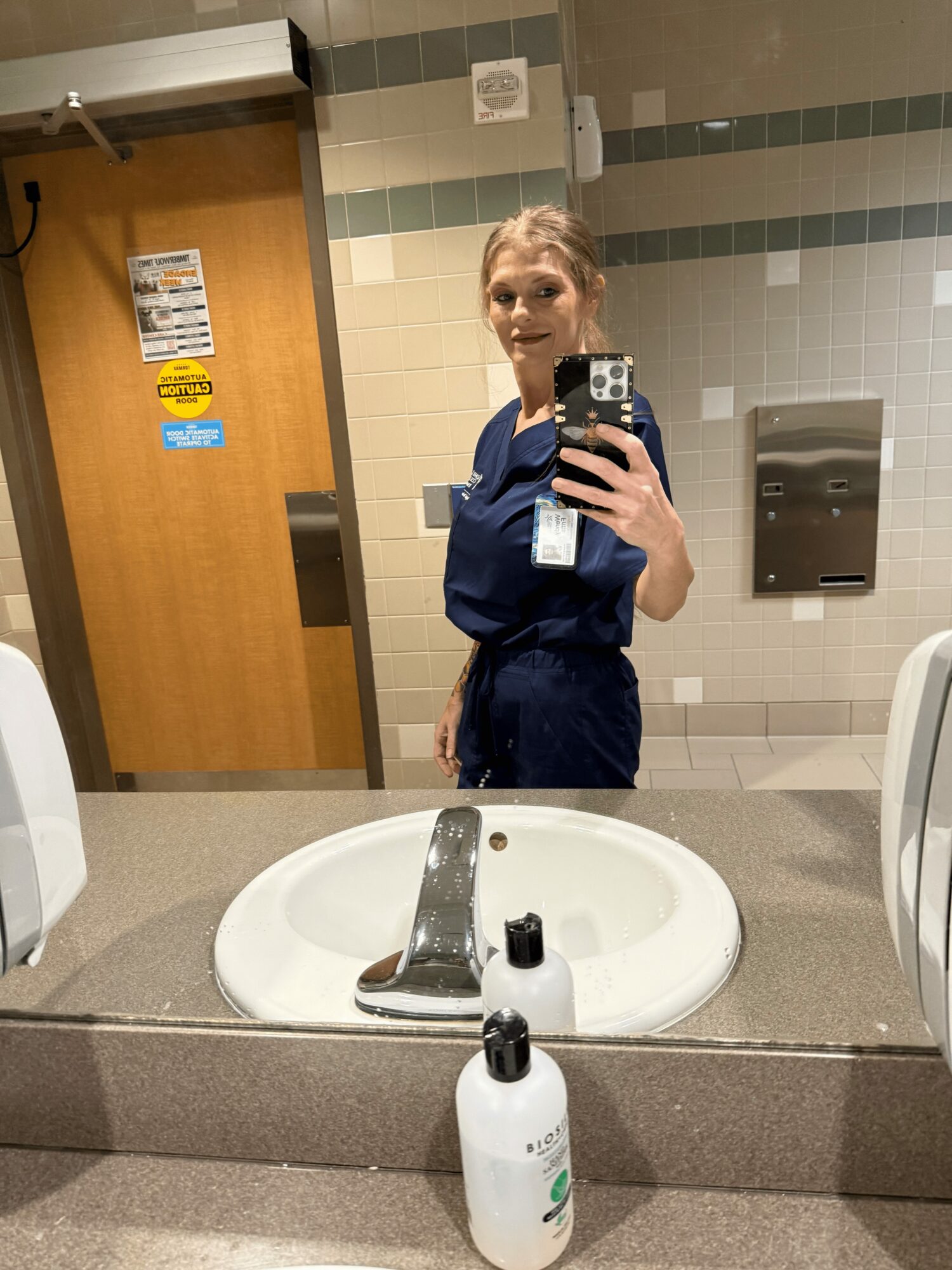Person taking a mirror selfie in a bathroom, wearing dark clothing, with a sink and soap dispenser visible.