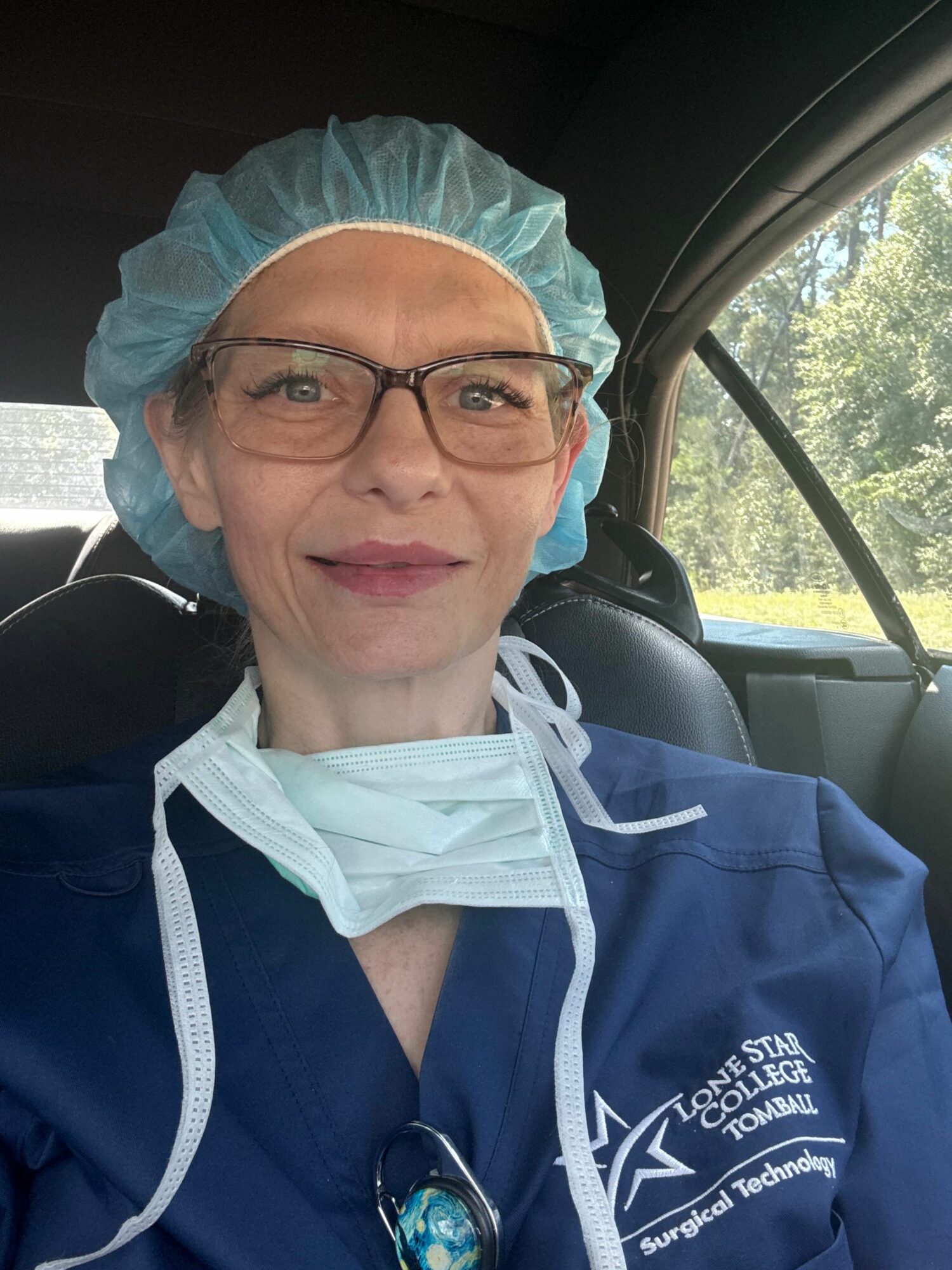 Person wearing glasses, a hair cap, and a face mask pulled down, sitting in a vehicle with trees outside.