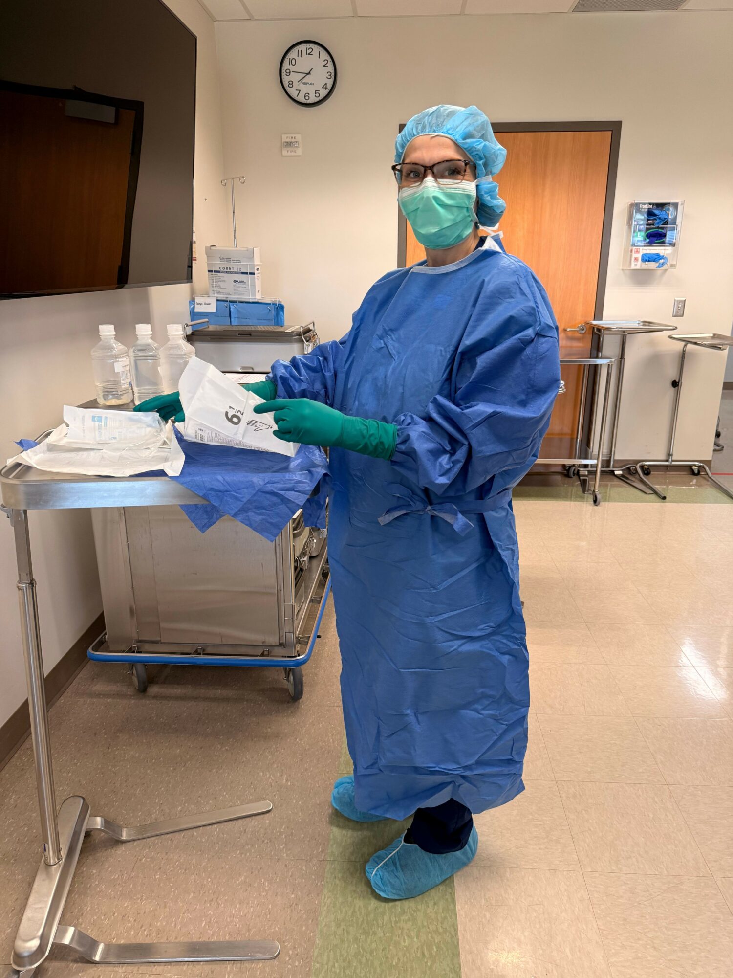 Healthcare worker in blue gown, mask, and gloves preparing supplies in a clinical setting.