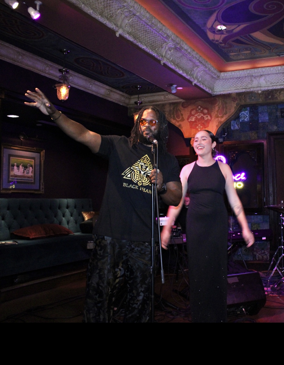 Man with glasses and beard speaking into microphone, woman smiling beside him, stage with colorful ceiling in background.