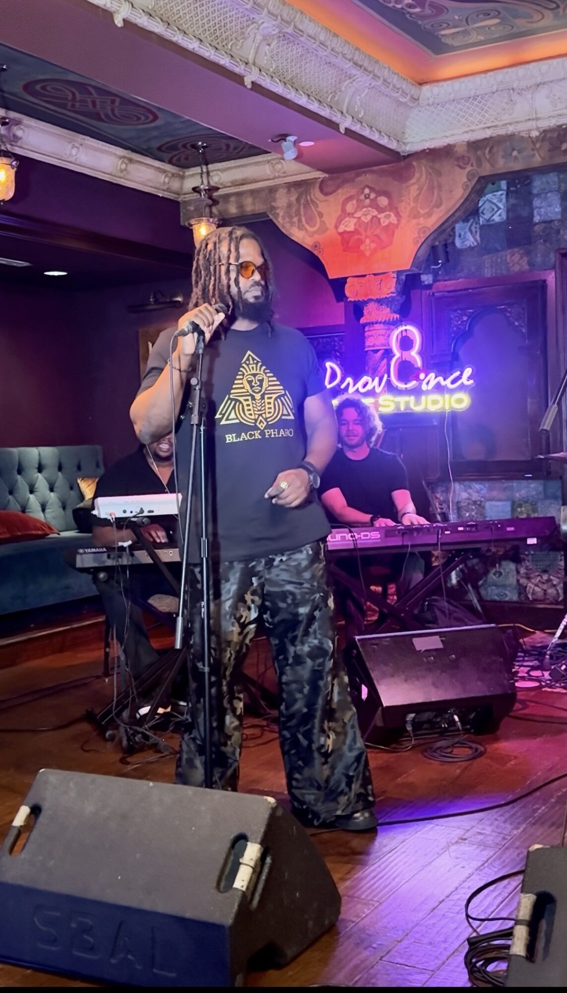 Performer with dreadlocks and face mask singing into microphone on stage, with DJ in background, colorful lighting, and sound equipment.