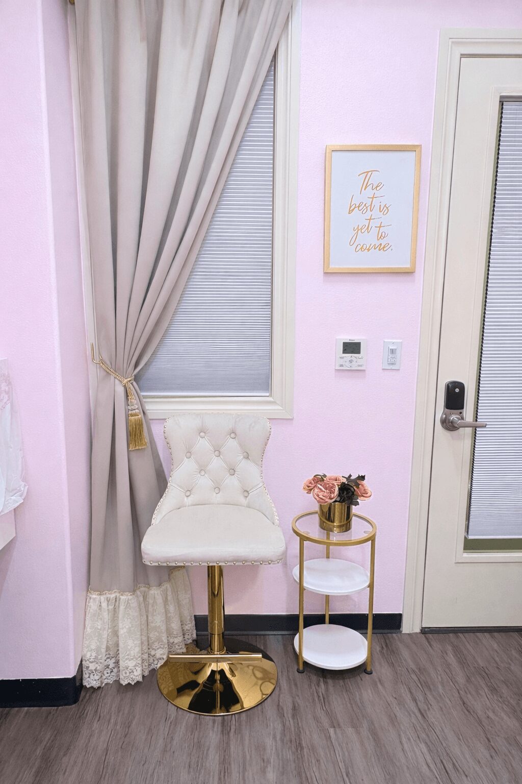 Corner with a white tufted chair, small round table with flowers, window with curtains, and framed quote on pink wall.