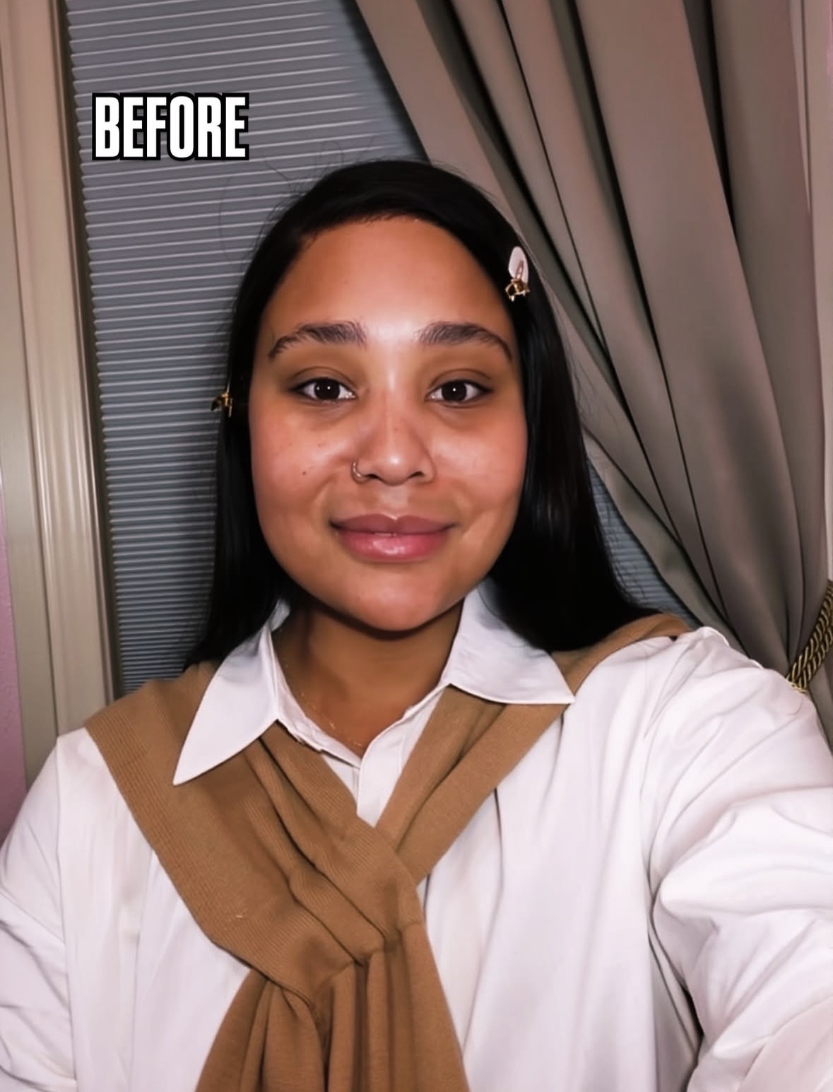 Young woman with long dark hair, wearing a white shirt and brown sweater, smiling indoors near curtains and window.
