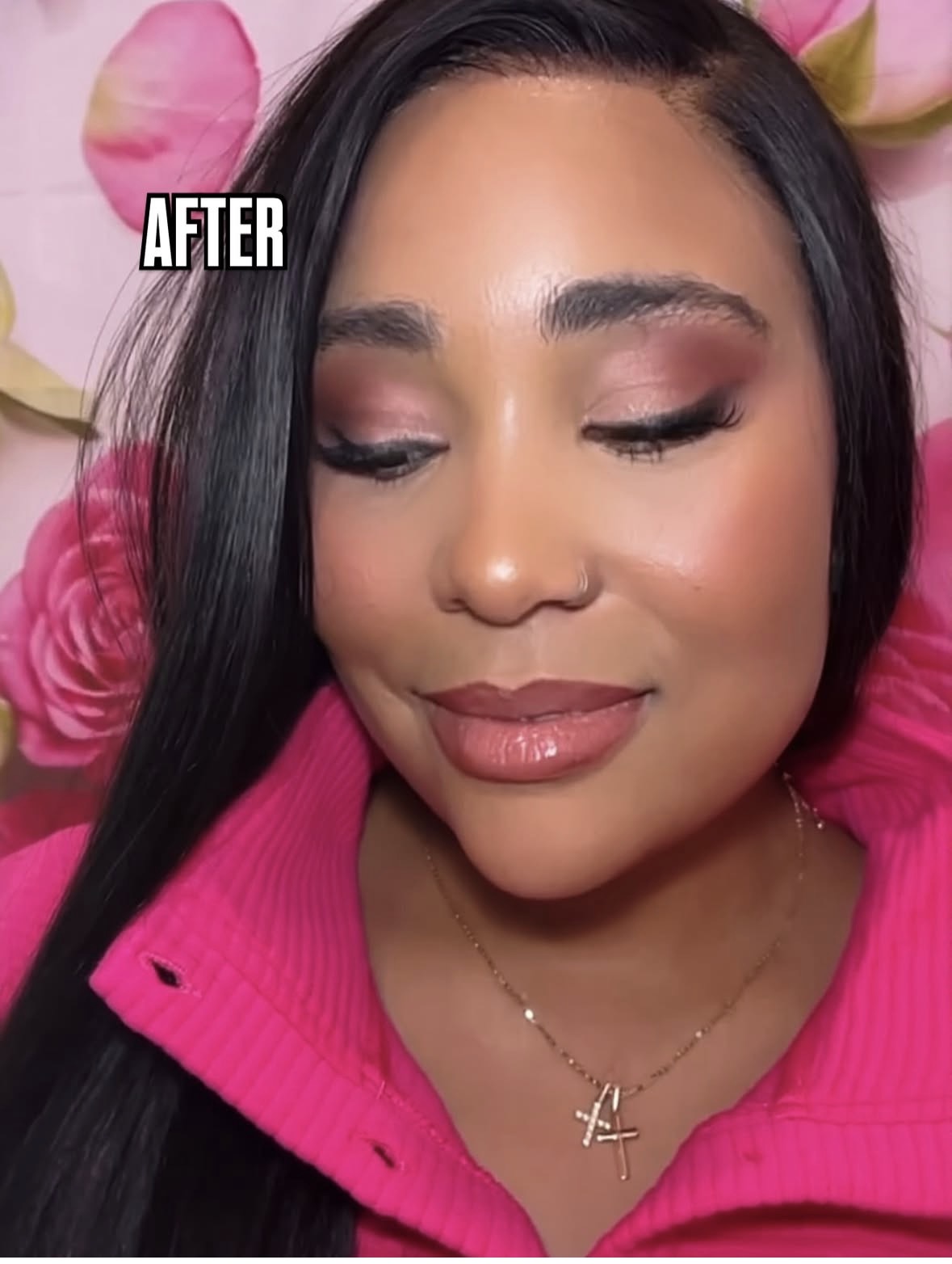Close-up of a woman with makeup, wearing a pink top, smiling with eyes closed, pink flowers in background.
