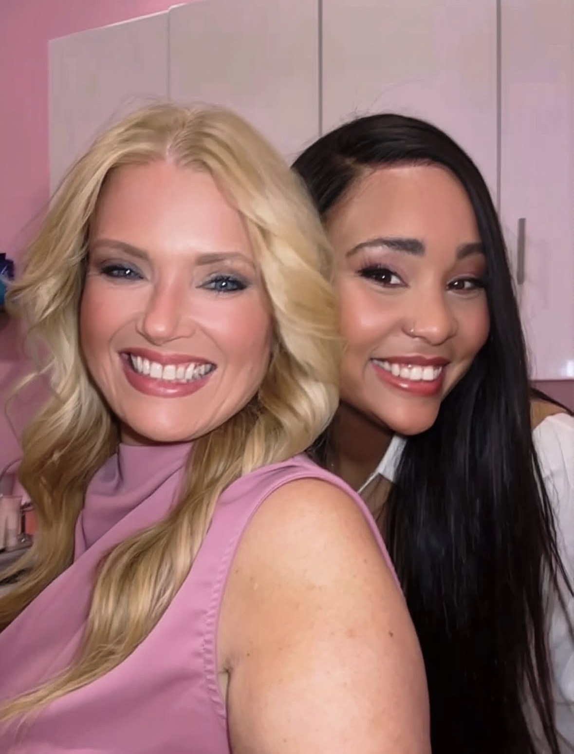 Two women smiling, one with blonde hair and the other with dark hair, in a room with pink and purple background.