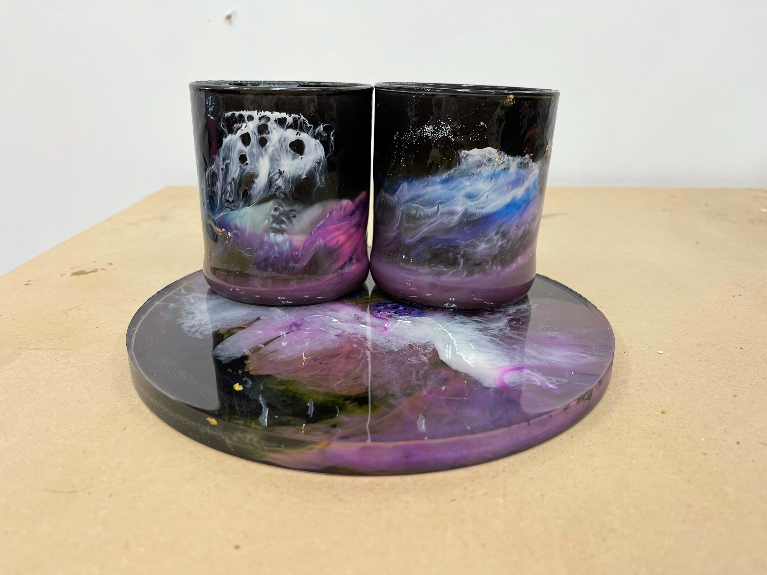Two black candles with colorful swirling patterns sit on a matching round tray with similar design, on a wooden surface.