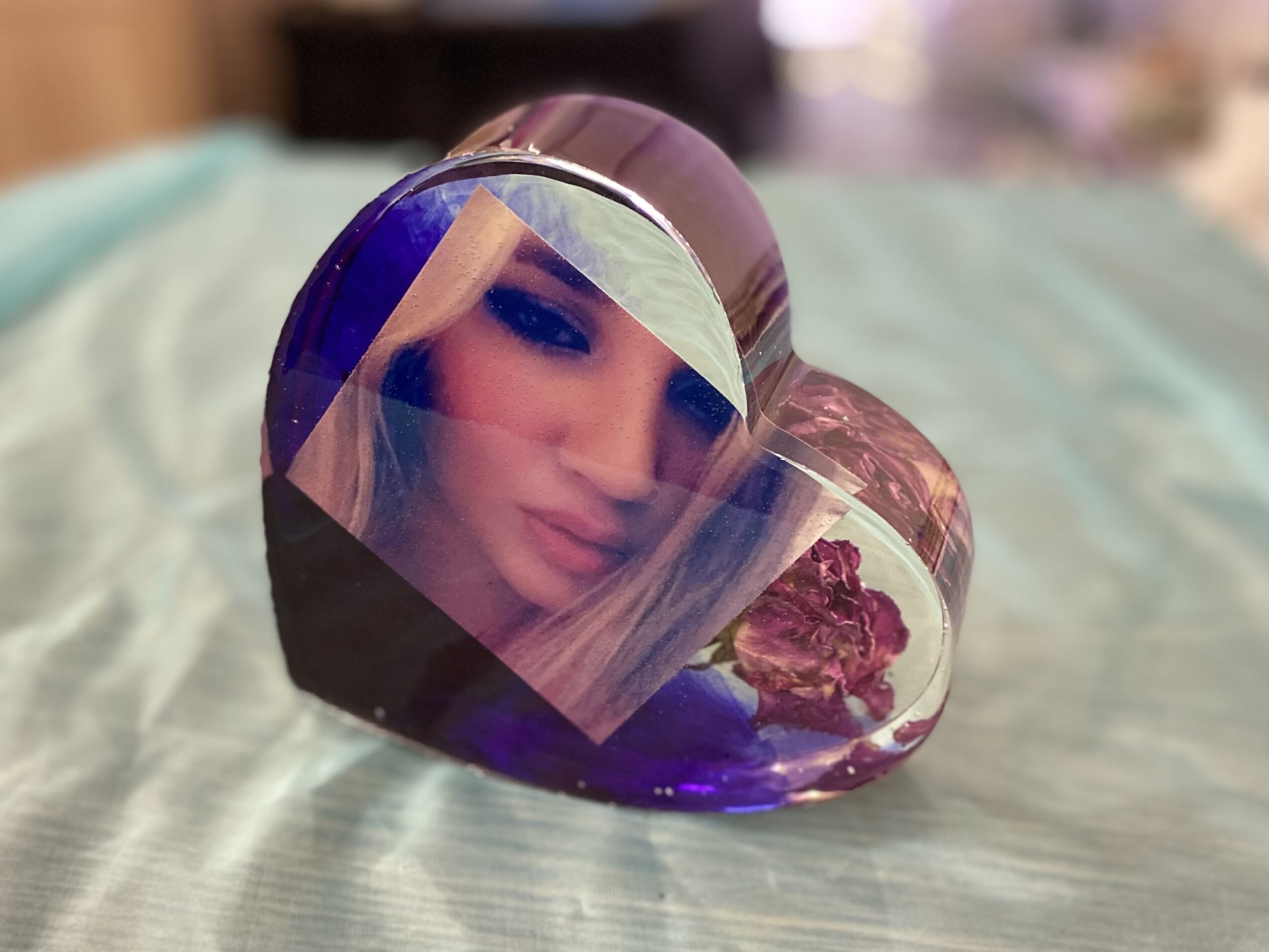 Heart-shaped glass container with purple and clear sections, reflecting a woman's face with makeup, resting on a fabric surface.
