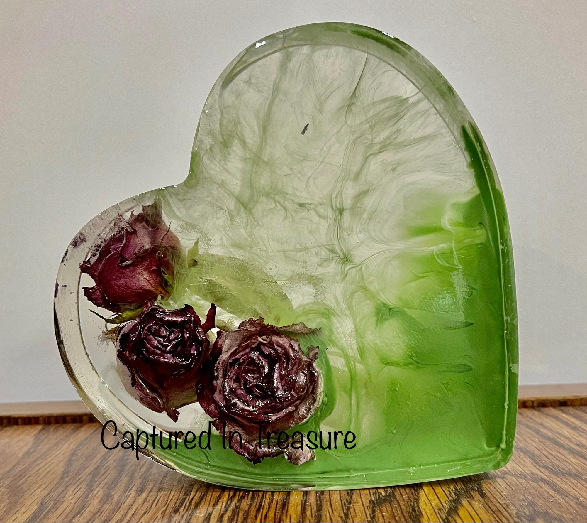 Heart-shaped glass container with three dried roses inside, partially filled with green liquid, on a wooden surface.