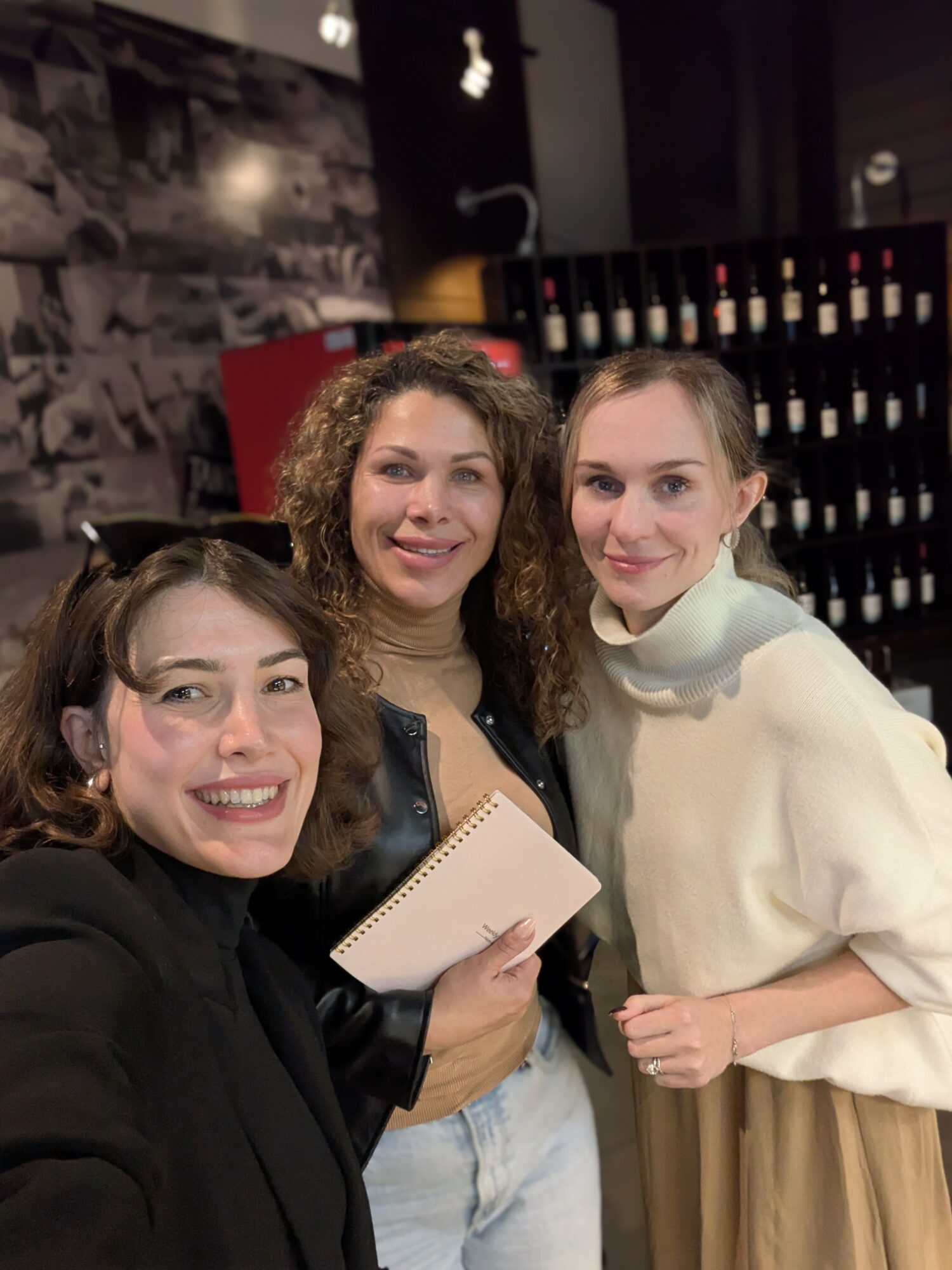 Three women smiling and posing for a photo indoors, with a wine rack in the background.