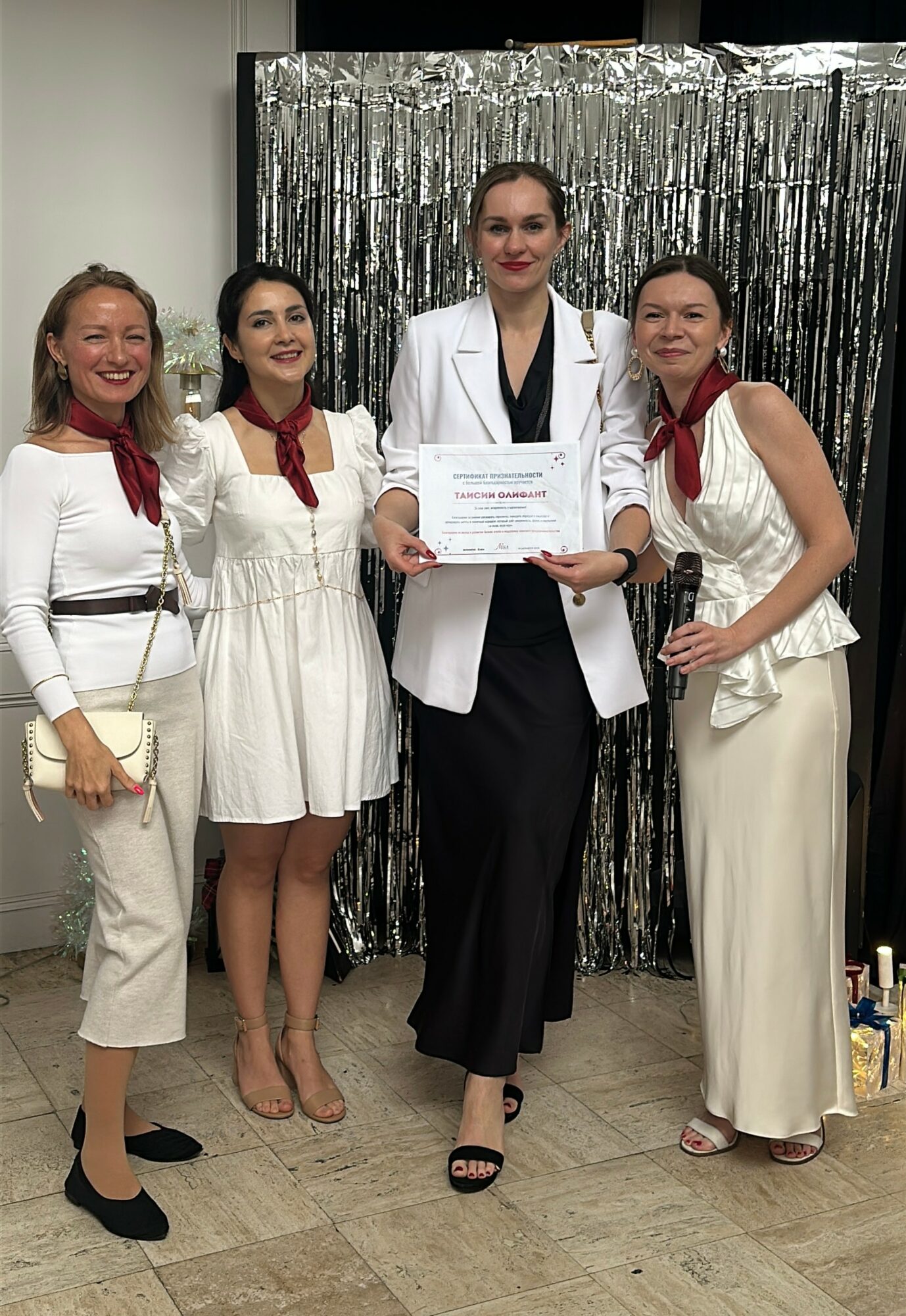 Four women standing together, one holding a certificate, in front of a silver curtain backdrop.
