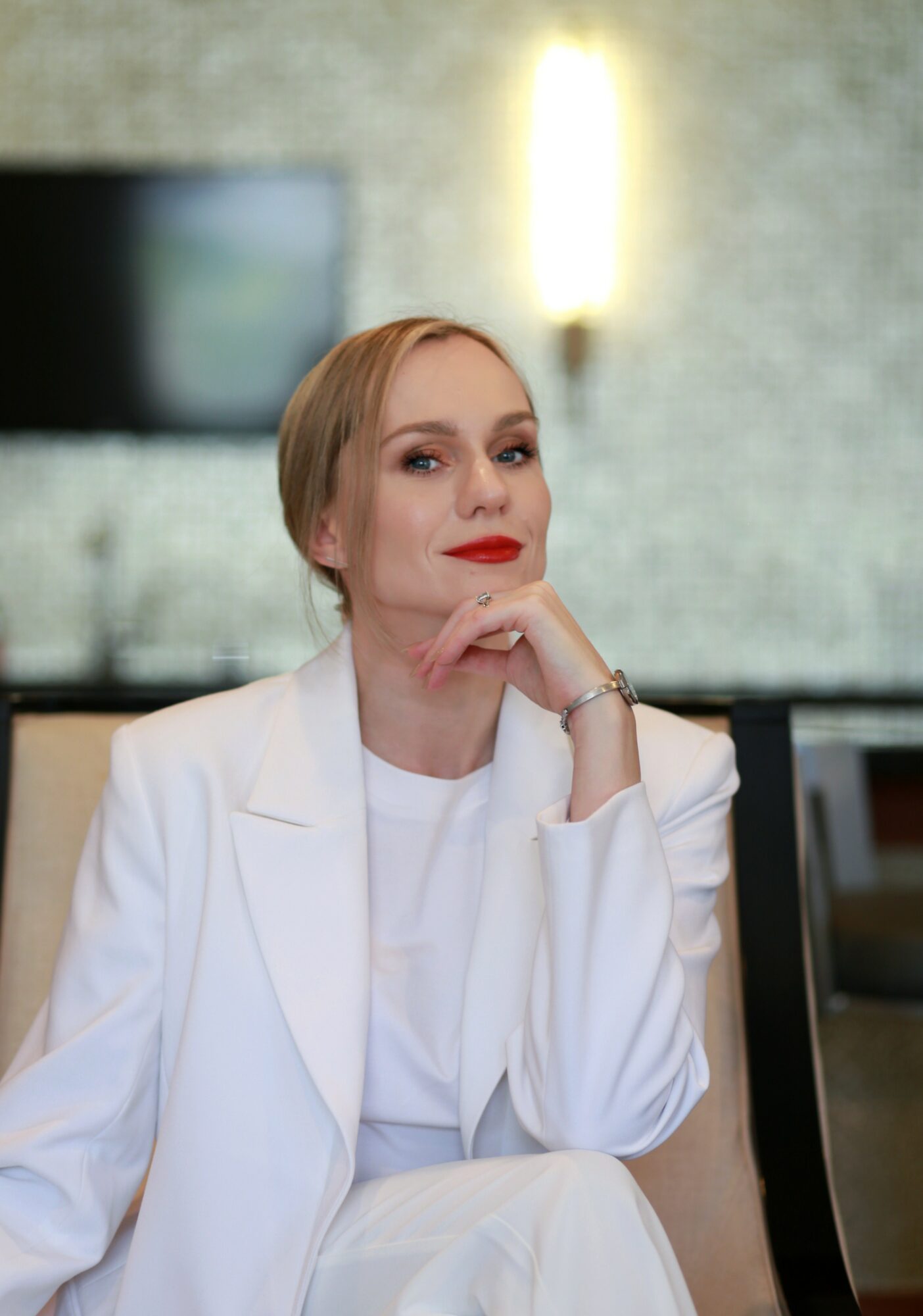 Woman with blonde hair and red lipstick sitting in a modern room, resting her chin on her hand, wearing a white blazer.