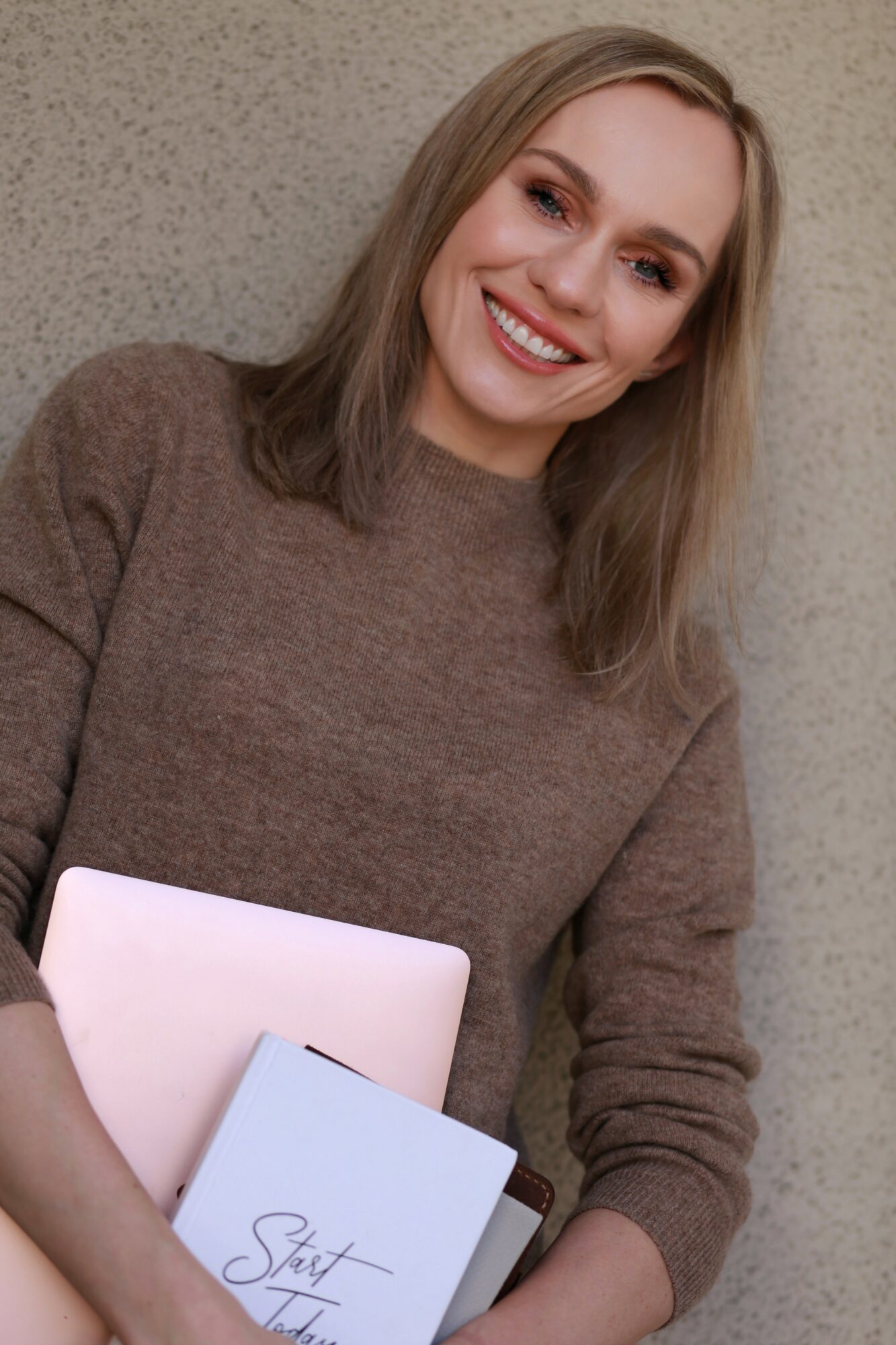 Young woman with shoulder-length hair smiling, holding notebooks, wearing a brown sweater, standing against a textured wall.