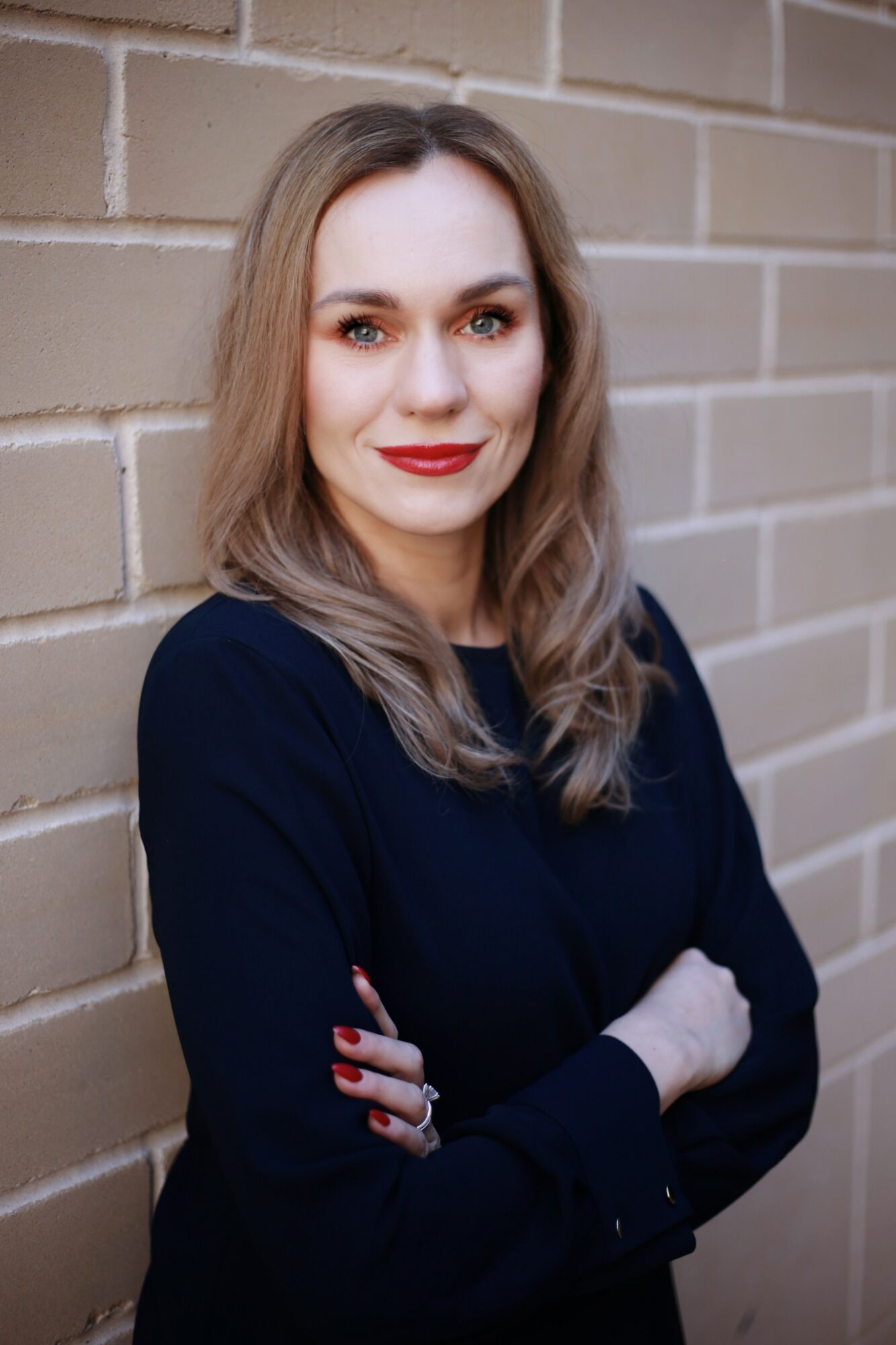 Woman with shoulder-length hair, red lipstick, wearing a dark top, standing against a brick wall, arms crossed.