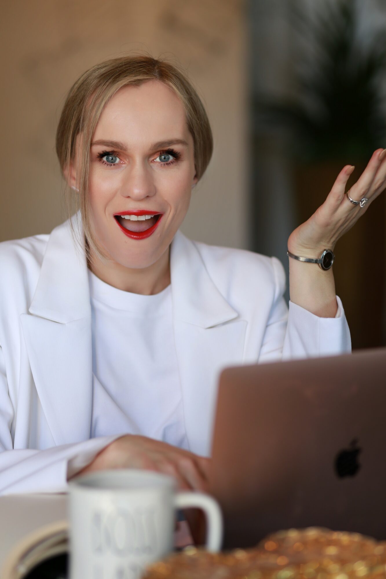 Woman with short blonde hair, white blazer, and red lipstick, gesturing with her right hand, sitting in front of a laptop.