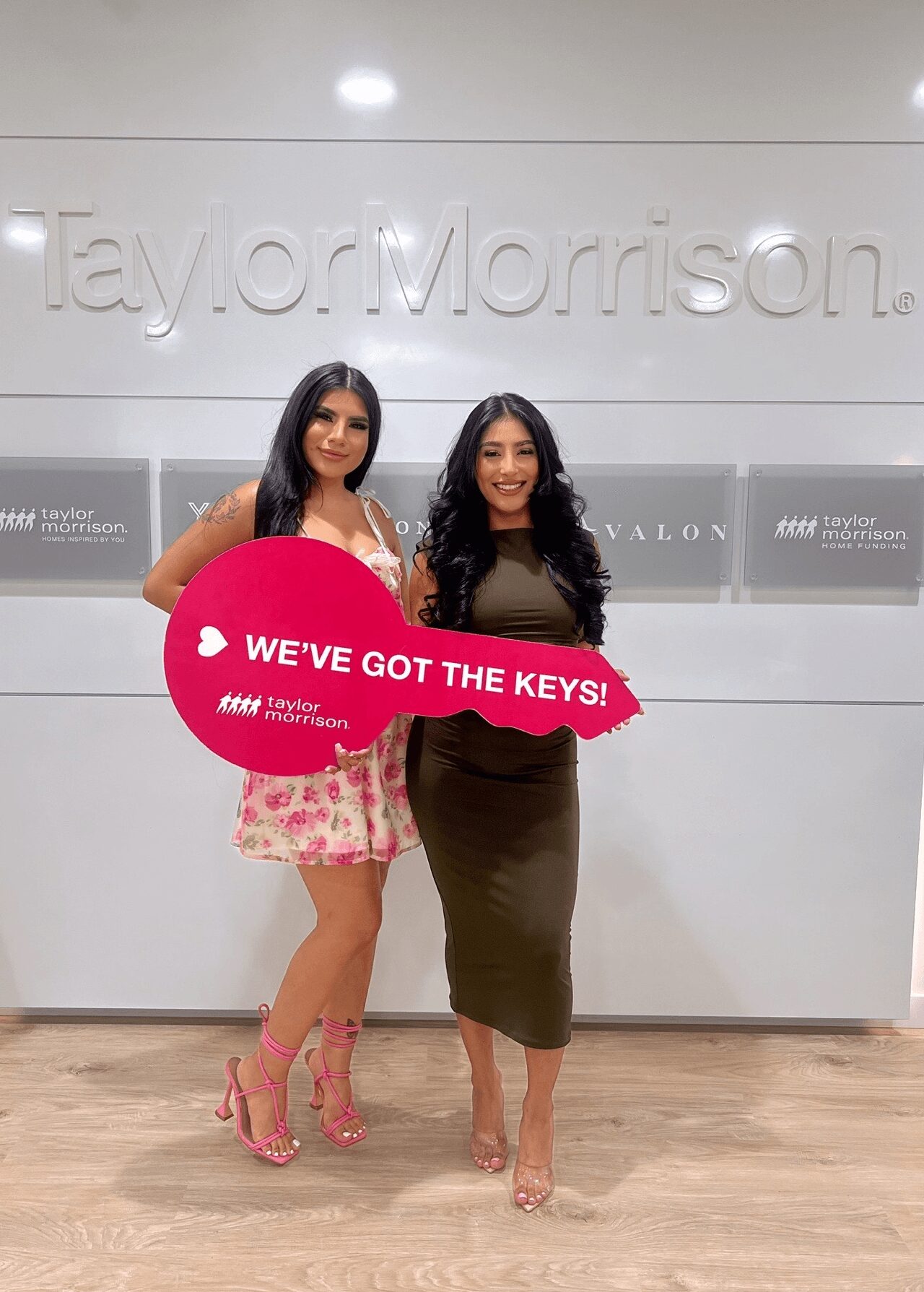 Two women standing indoors holding a pink sign that says 'WE'VE GOT THE KEYS!'.