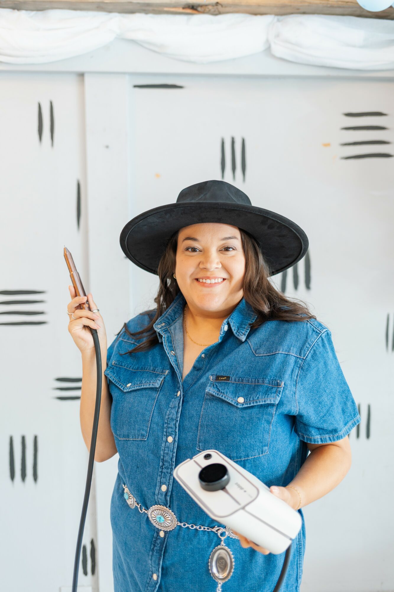 Young woman in denim shirt and black hat smiling, holding a cleaning tool and a spray bottle, standing in front of lockers.