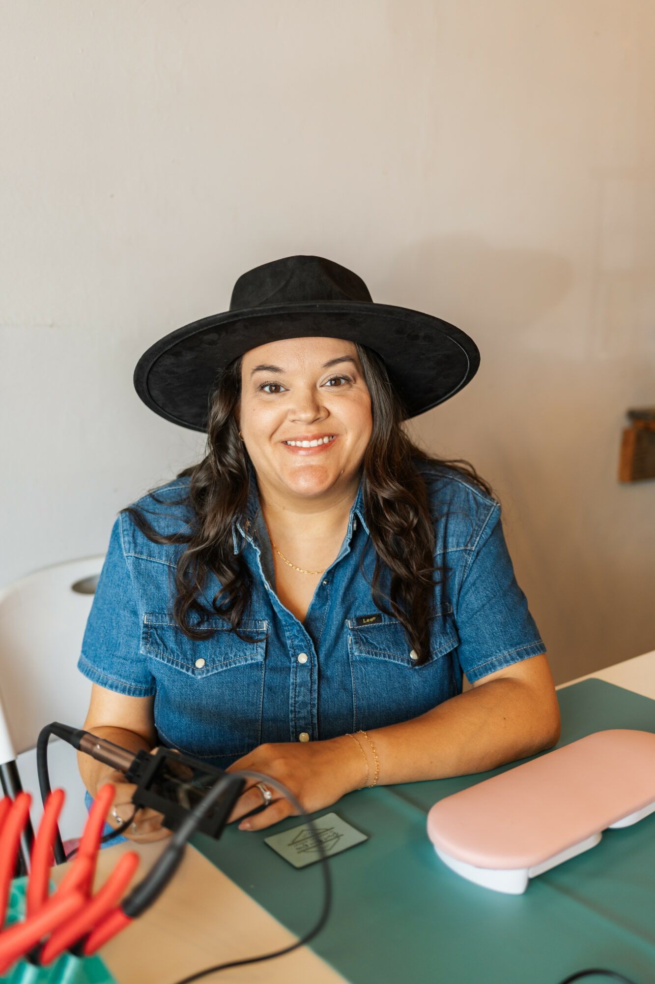 Woman with long dark hair wearing a wide-brimmed hat and denim shirt, smiling, sitting at a table with various objects.