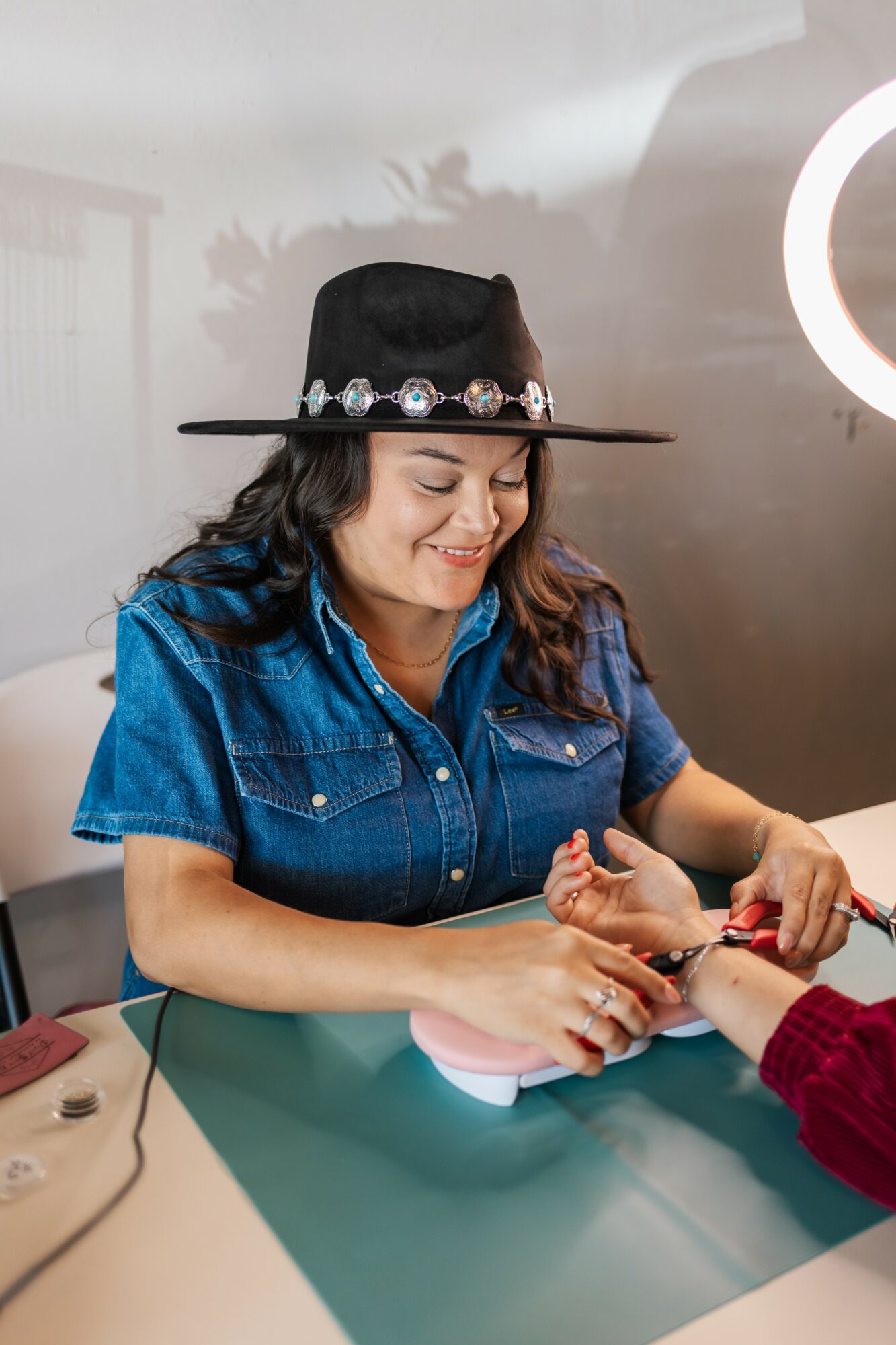 Woman wearing a black hat and denim shirt, smiling while receiving a manicure, seated at a table.