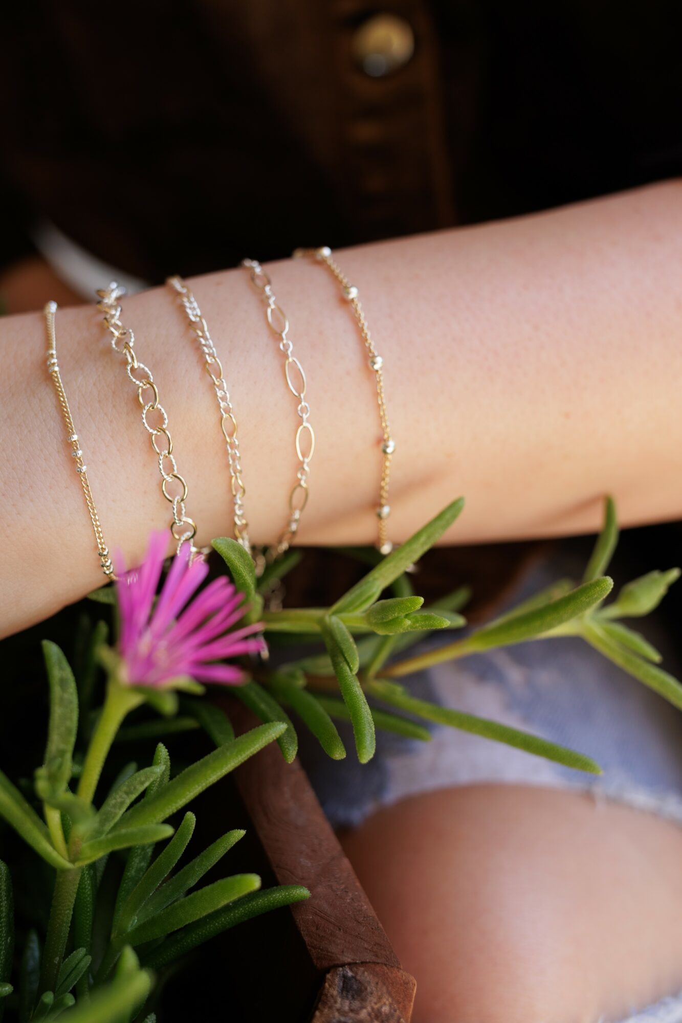 Person's arm with four gold bracelets resting on a green plant with pink flowers, wearing denim shorts.