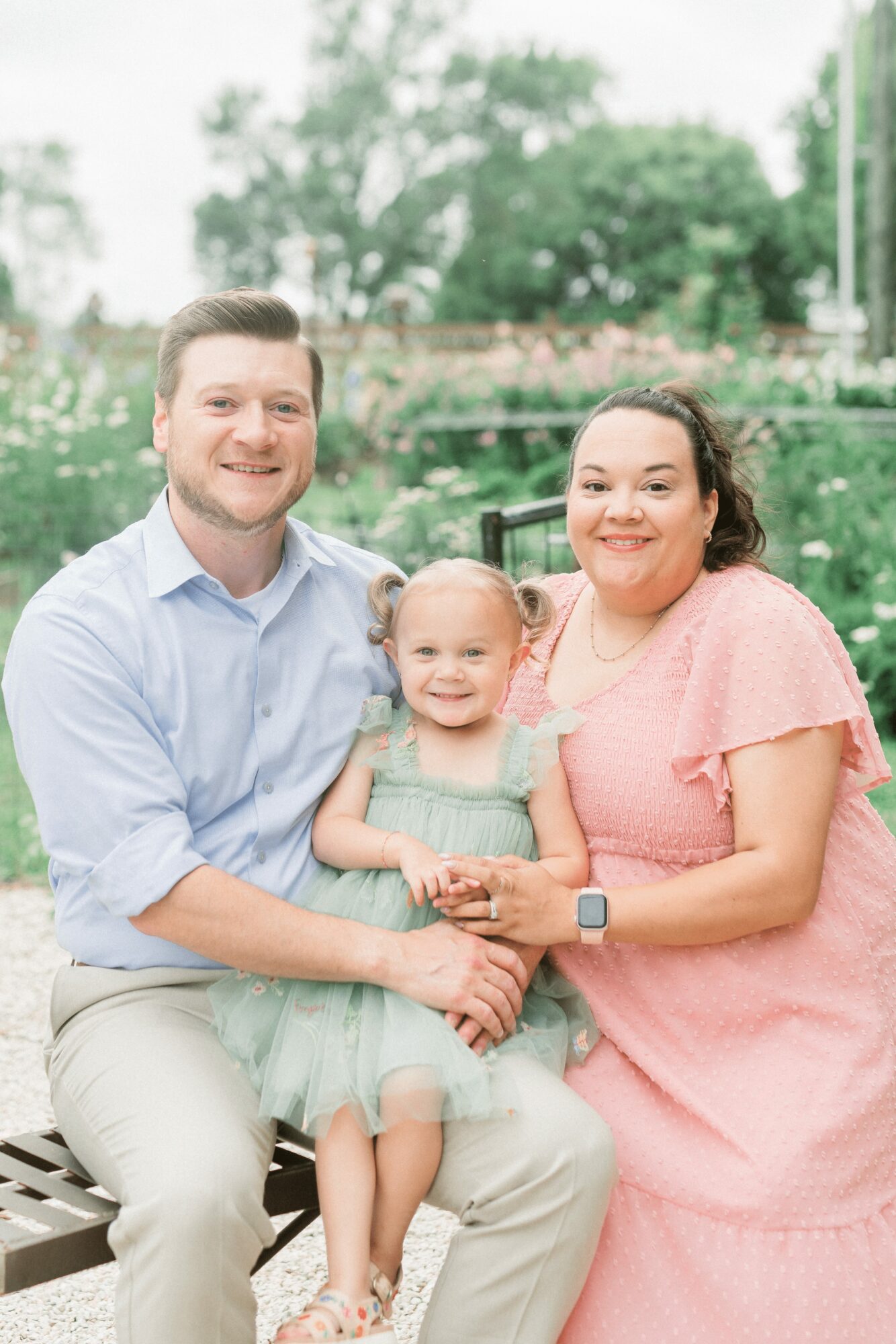 Family of three sitting outdoors, smiling, with trees and water in background, woman in pink dress, man in light shirt, young girl in dress
