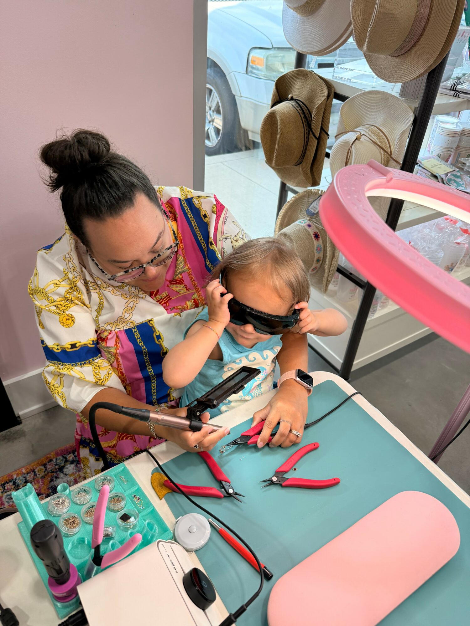 Adult and child wearing sunglasses, sitting at a table with tools and accessories, hats visible outside window.